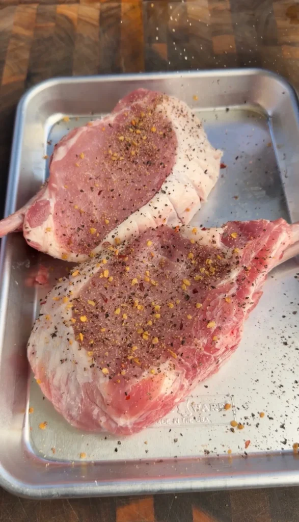 Two raw double bone pork chops seasoned with herbs and spices on a metal baking tray ready for cooking