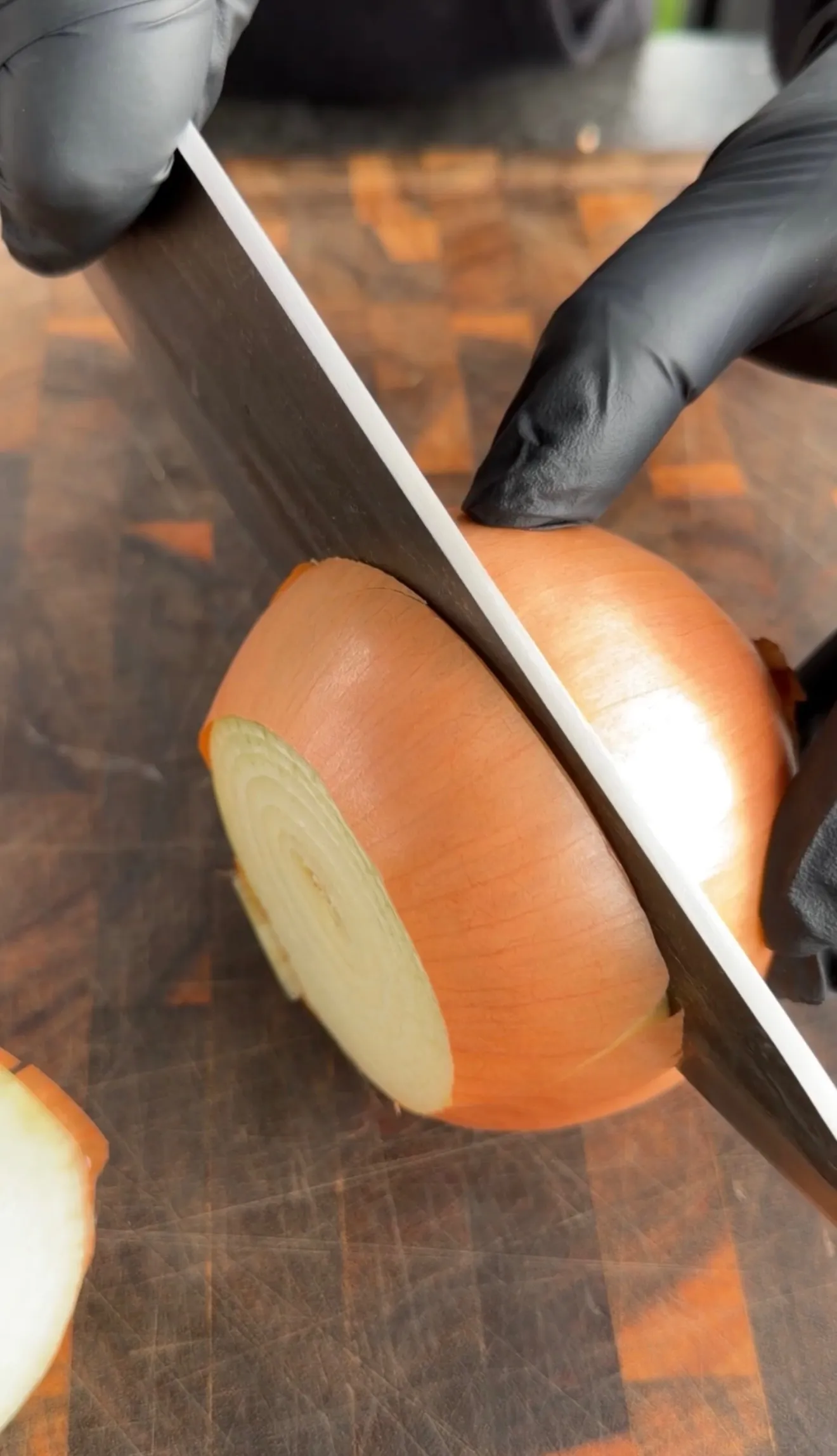 Hands in black gloves slicing a large onion with a knife on a wooden cutting board for crab stuffed bacon-wrapped rings