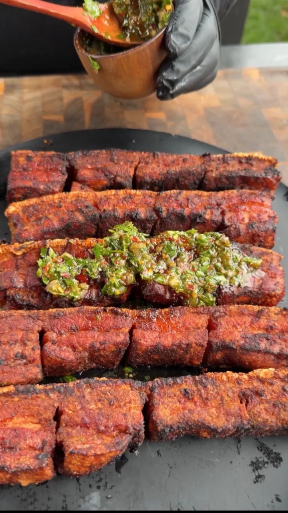 Sliced smoked and fried pork belly with green chimichurri sauce being spooned on top on a dark serving board