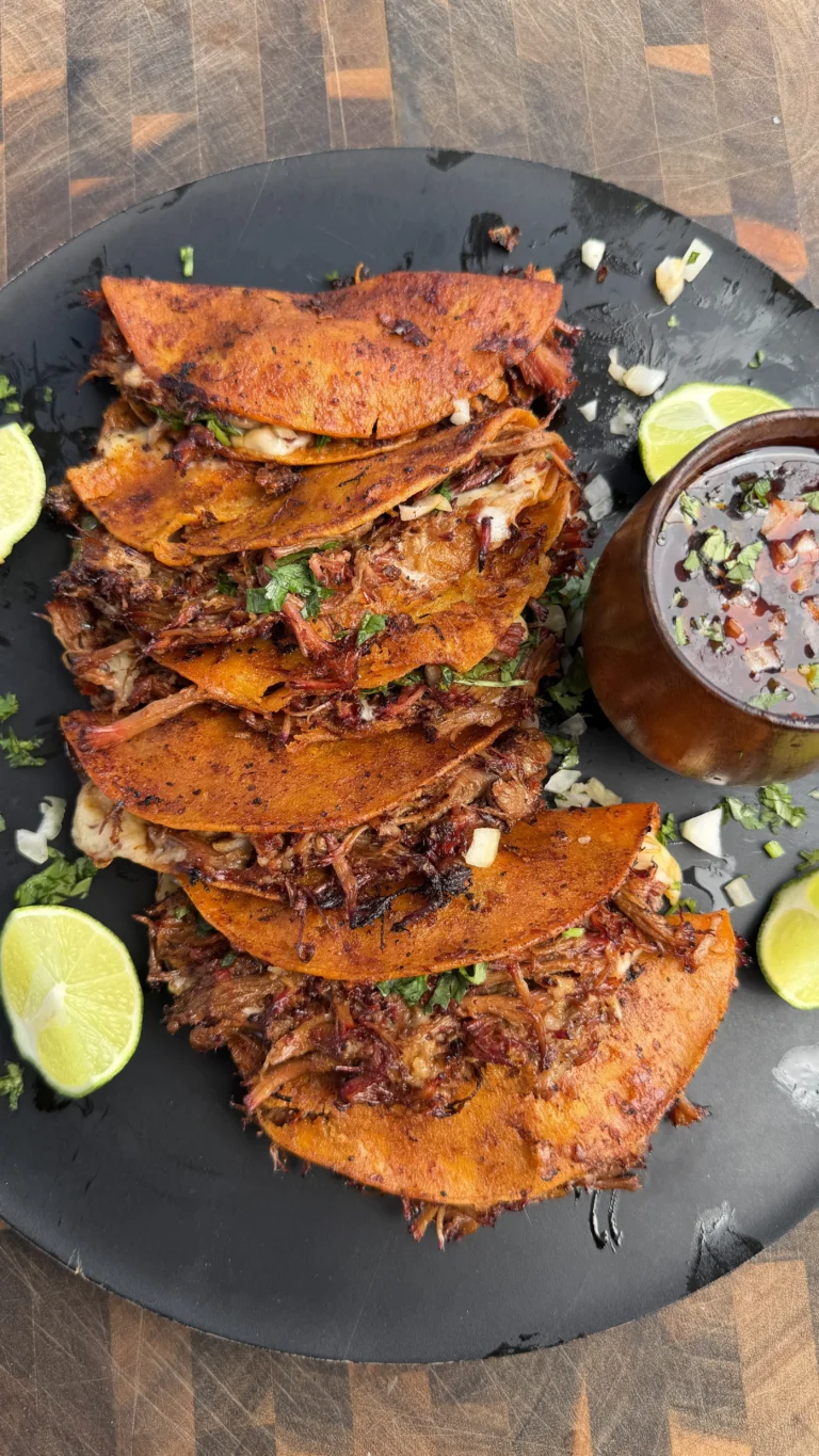 Stack of crispy smoked quesa birria tacos on black plate with lime wedges and consommé dipping sauce