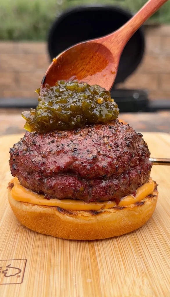 Spicy Juicy Lucy burger with melted cheese and green jalapeño relish being spooned on top on wooden board