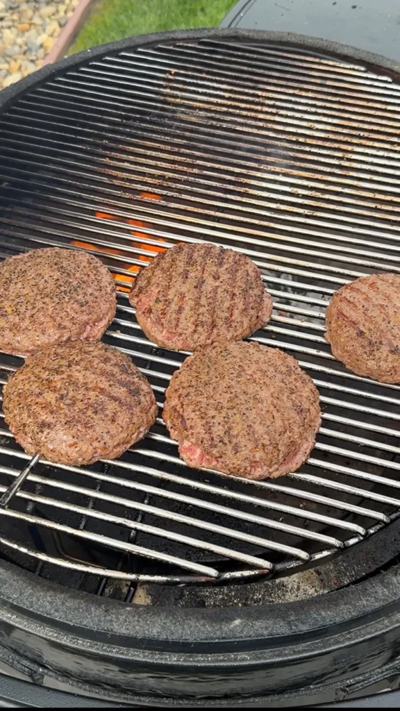 Five spicy Juicy Lucy burger patties cooking on a round grill grate with flames visible beneath