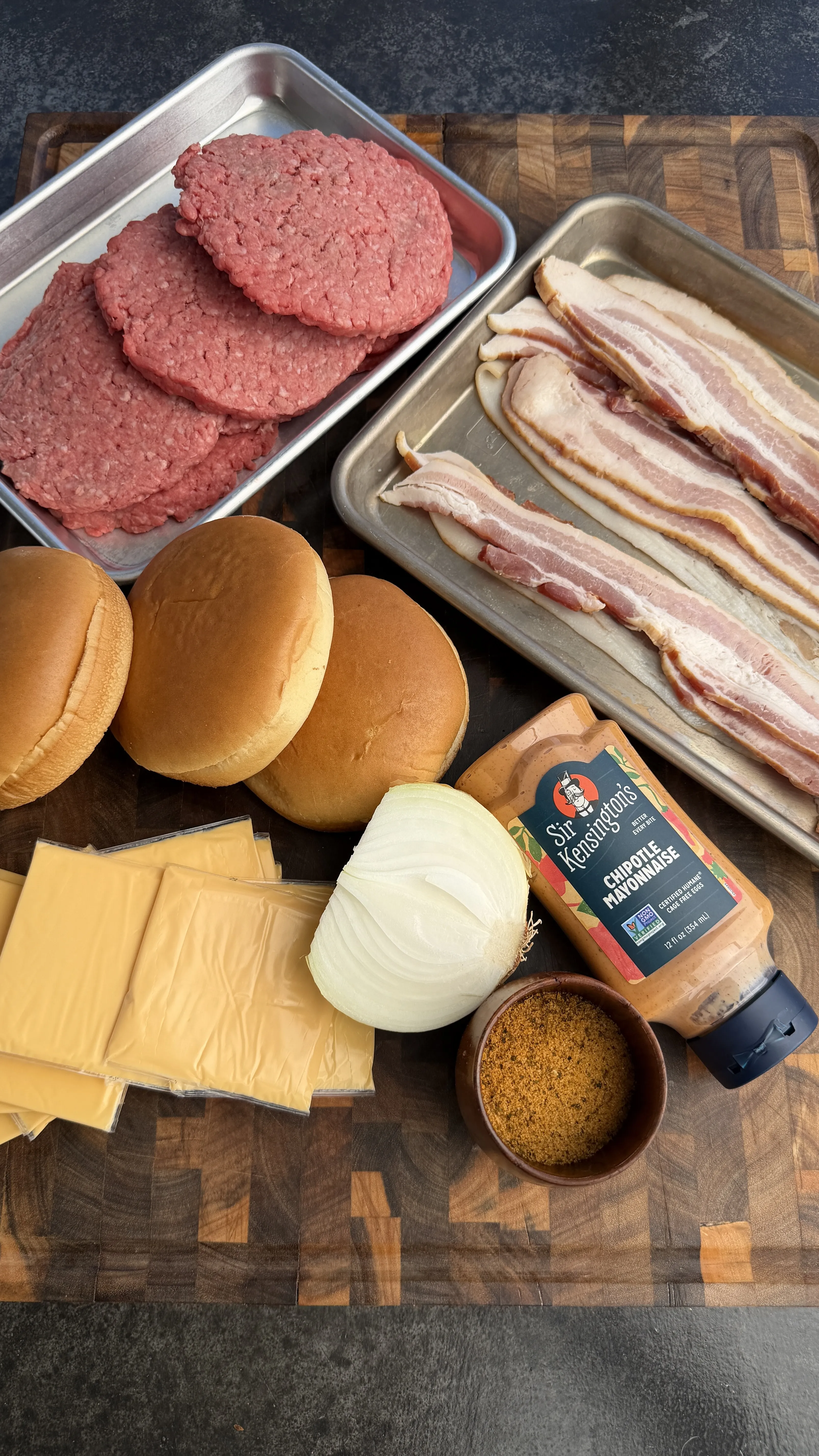 Raw burger patties, bacon strips, cheese slices, onions, and hamburger buns arranged on cutting board for patty melt