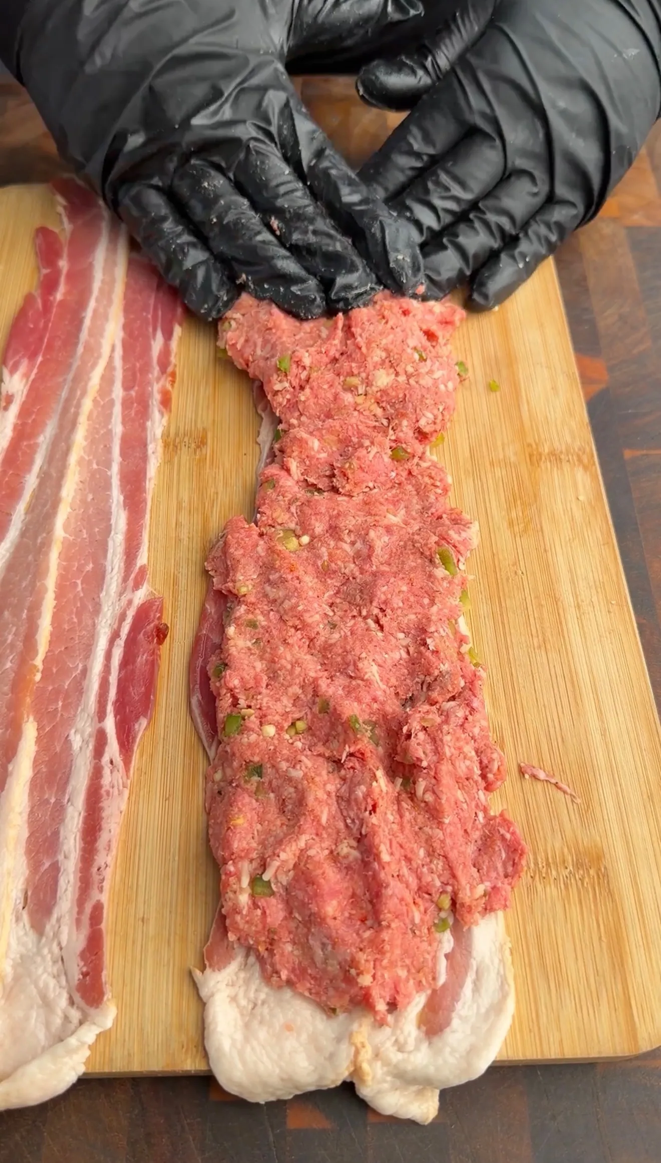 Raw ground beef mixture with jalapeños being shaped on bacon strips on wooden cutting board for skewer preparation