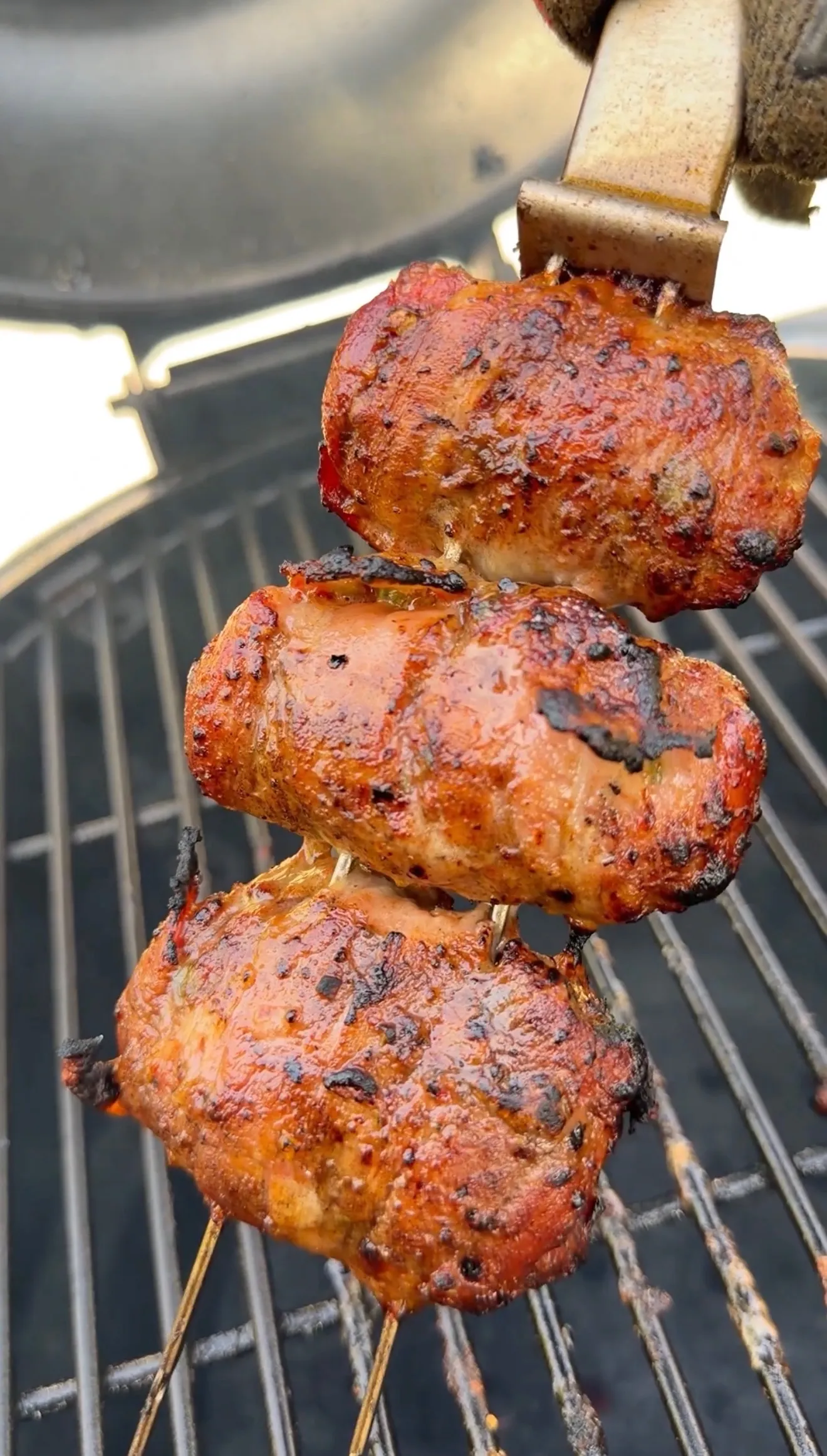 Three bacon-wrapped jalapeño beef skewers grilling on barbecue grates with golden-brown char marks