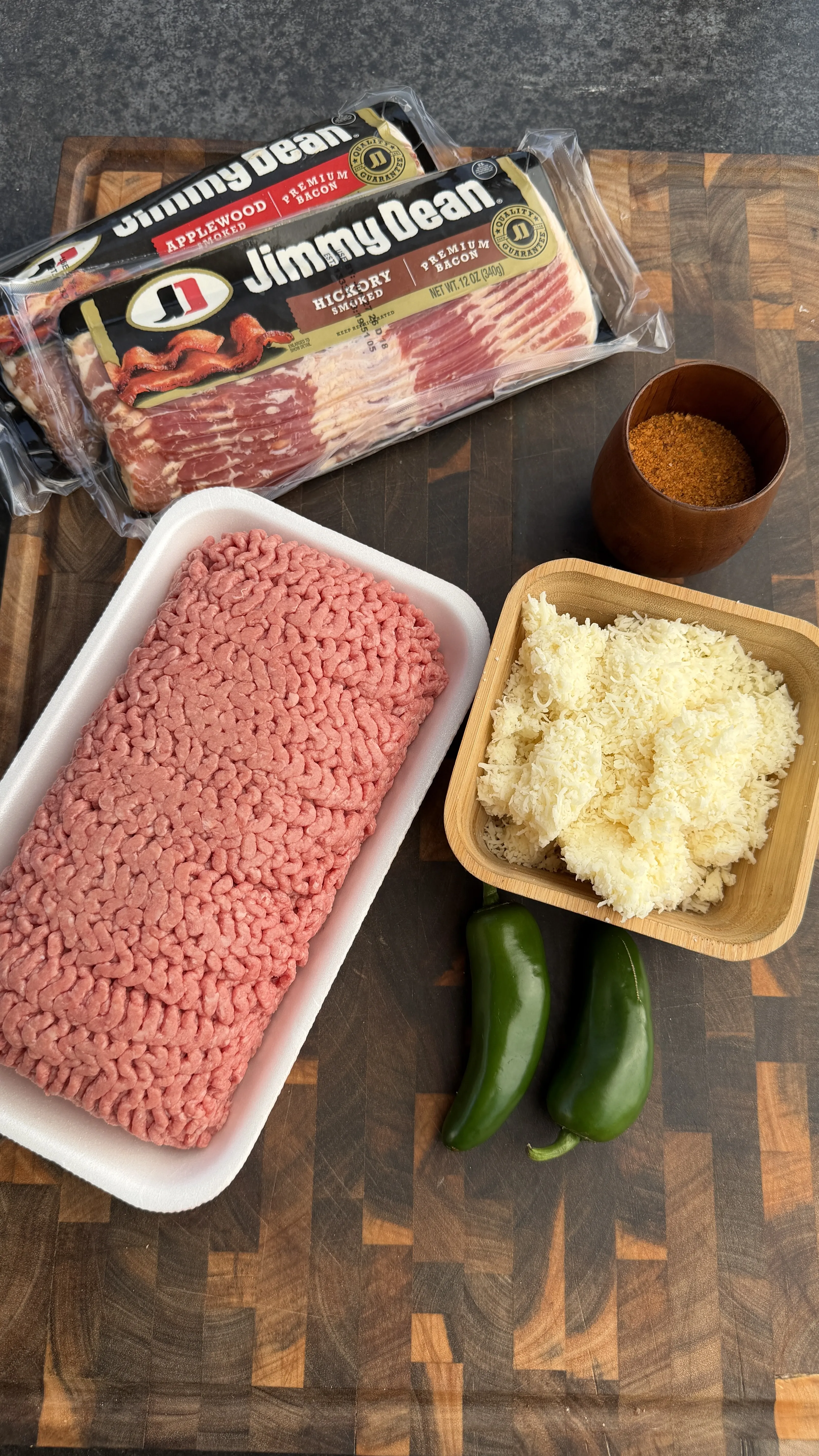Raw ground beef, Jimmy Dean bacon, shredded cheese, jalapeños and spices on wooden cutting board for skewer prep