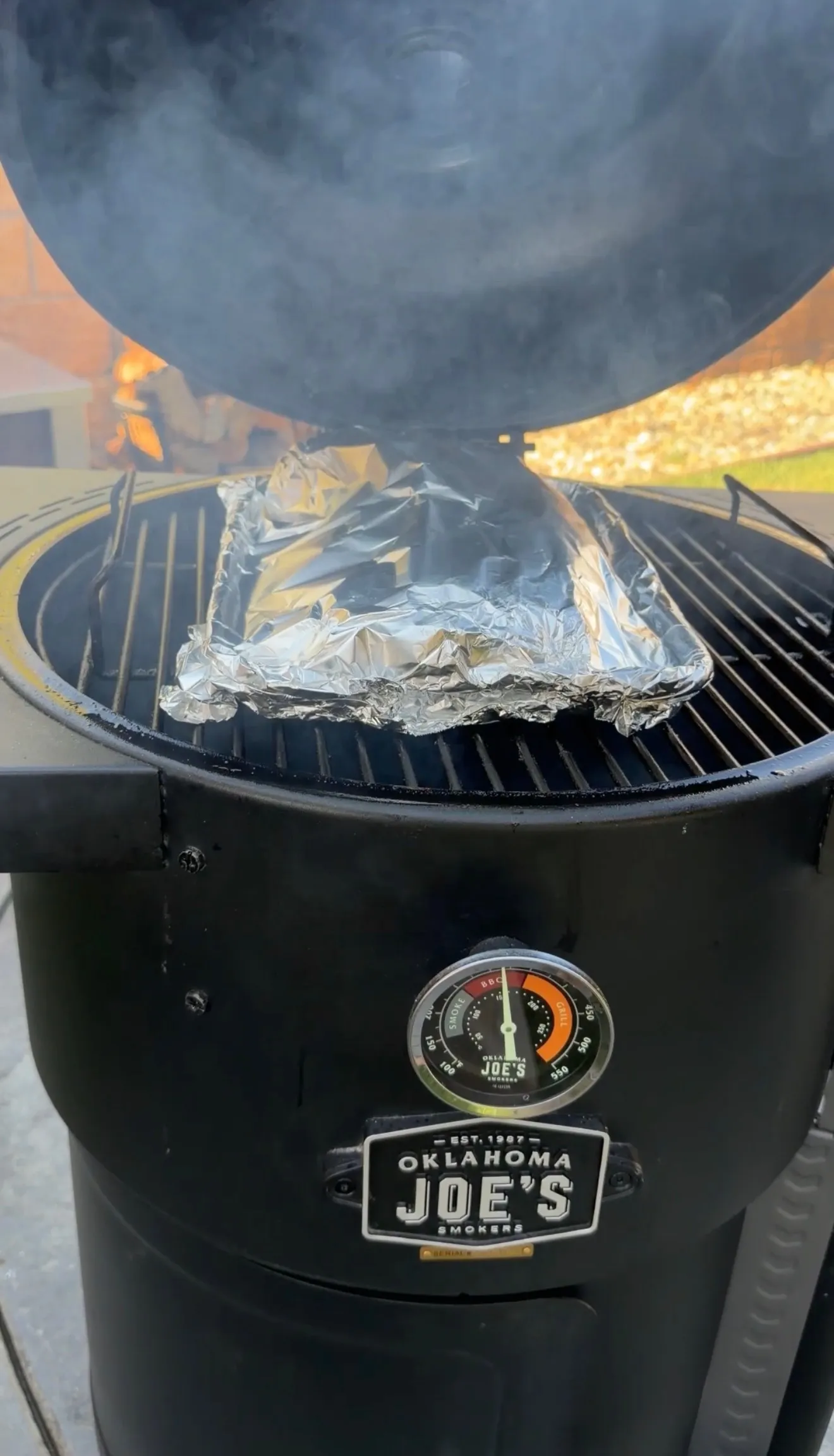 Beef ribs wrapped in aluminum foil smoking on Oklahoma Joe's smoker with temperature gauge visible