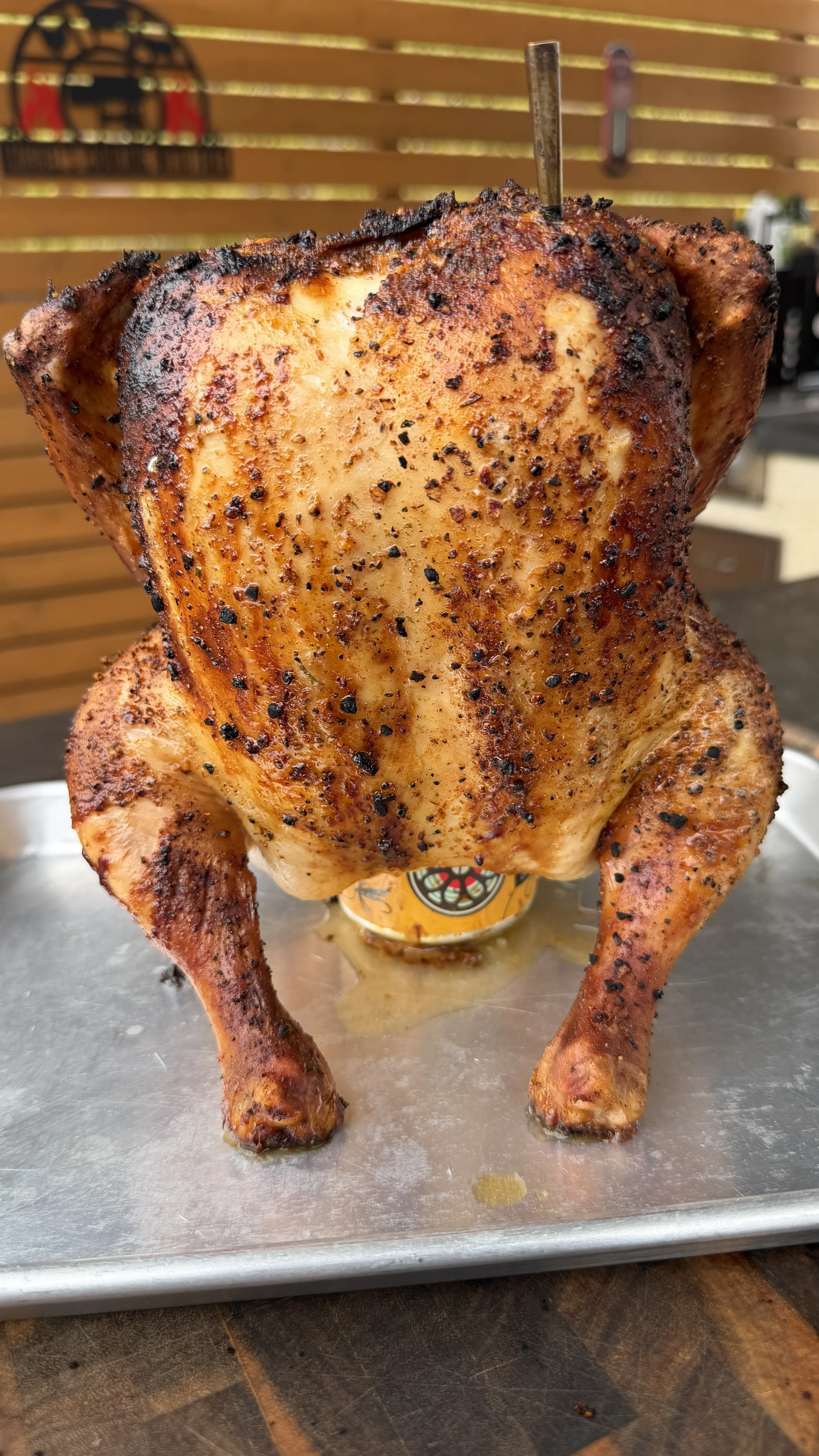 Golden brown beer can chicken standing upright on a beer can, seasoned with black pepper on a baking sheet