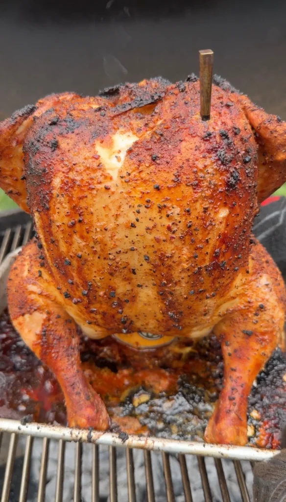 Beer can chicken with golden crispy skin and seasoning cooking upright on a barbecue grill grate