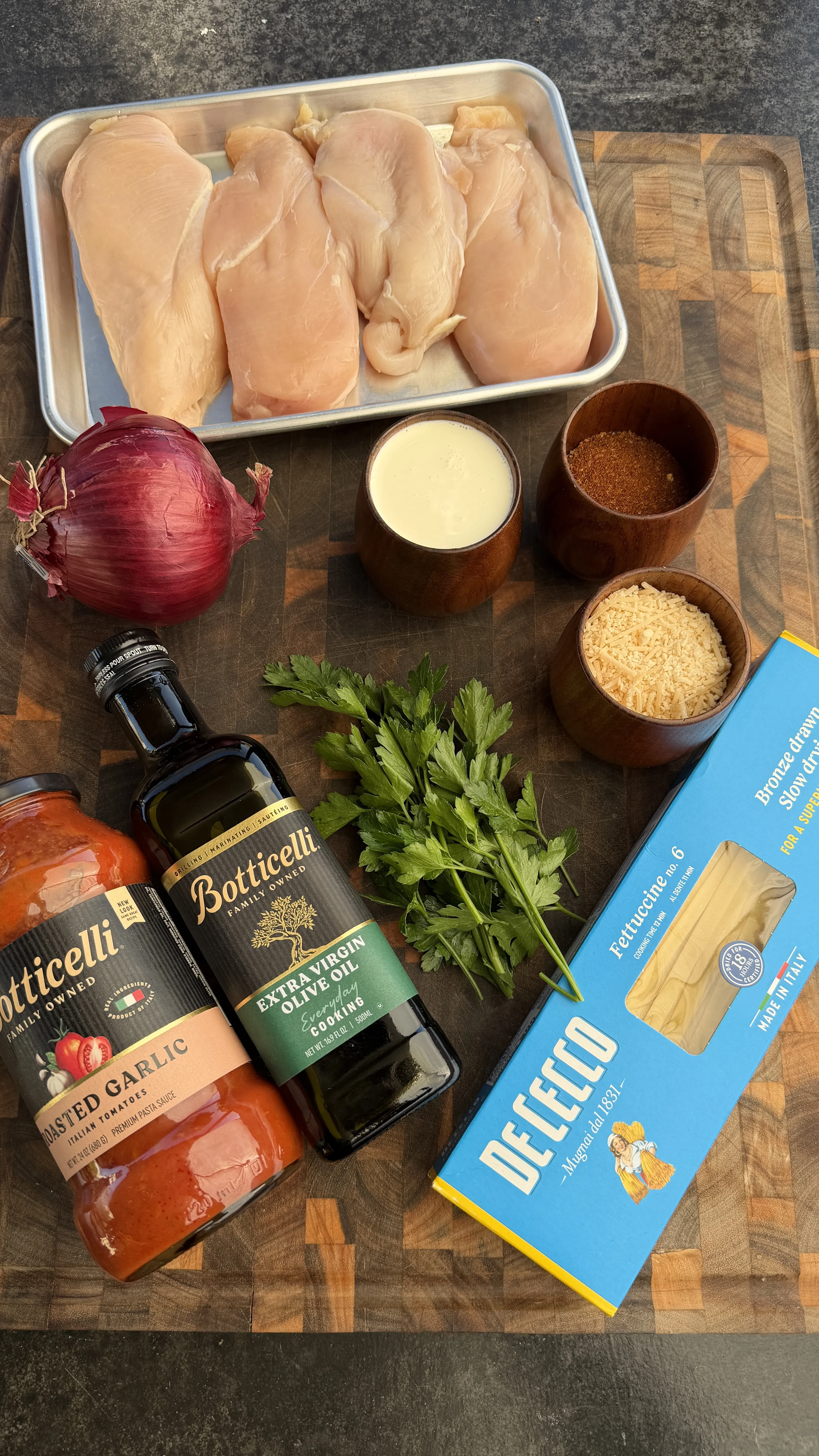 Cajun chicken fettuccine ingredients laid out on wooden cutting board including raw chicken, pasta, and spices