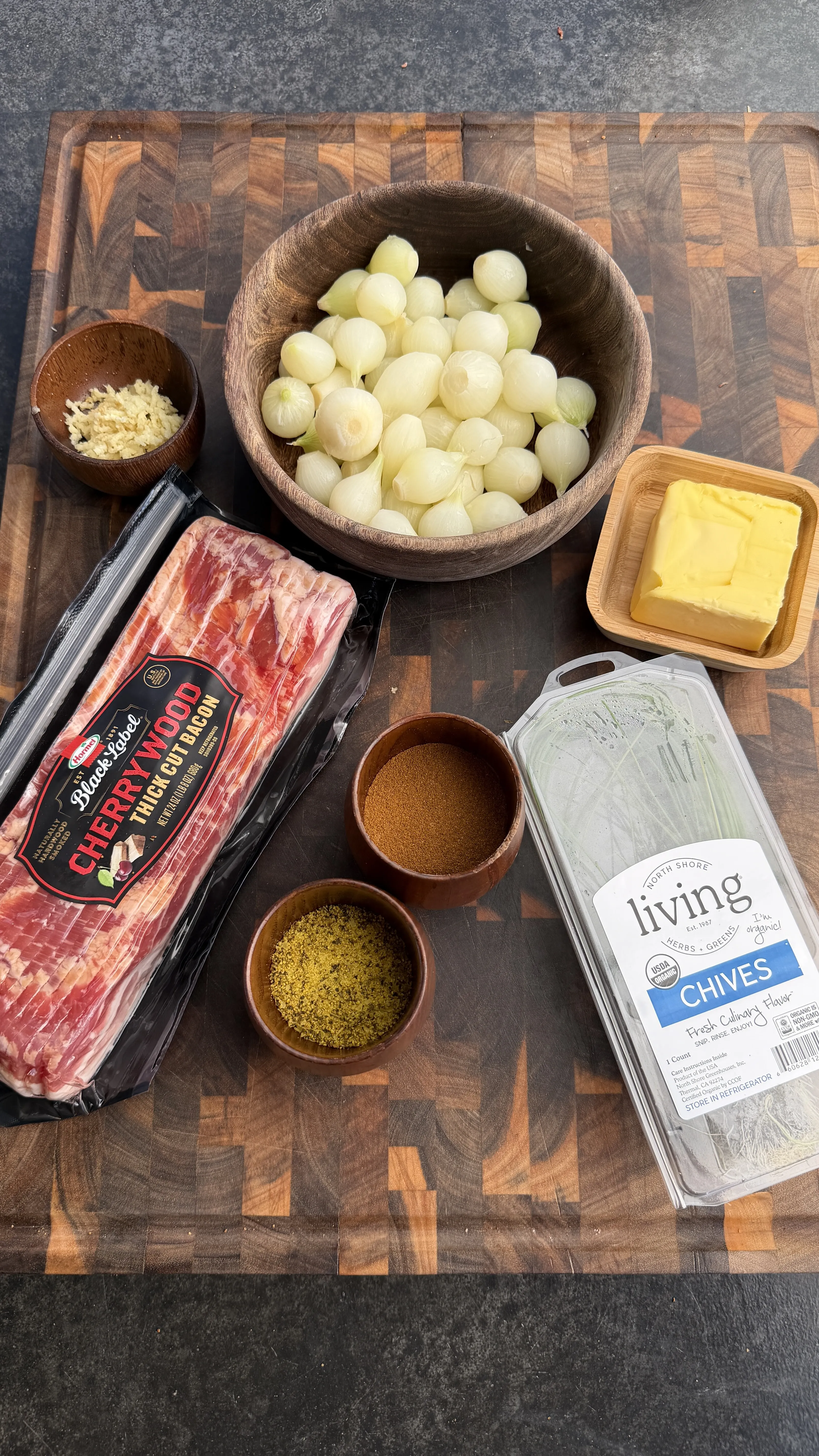 Mini Cajun onion boil ingredients on wooden cutting board: pearl onions, bacon, butter, chives, and spices