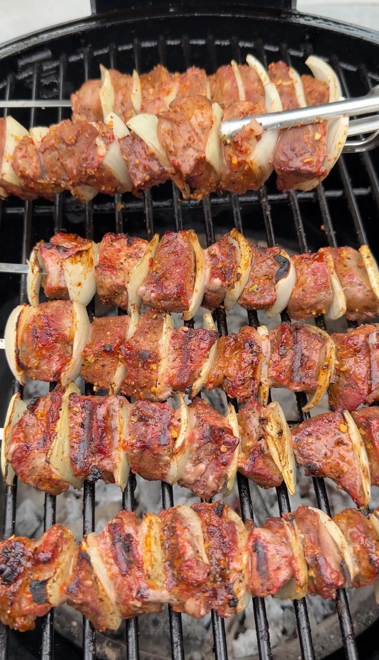 Cheese-crusted chimichurri steak kebabs with onions grilling on a round barbecue grill with metal tongs