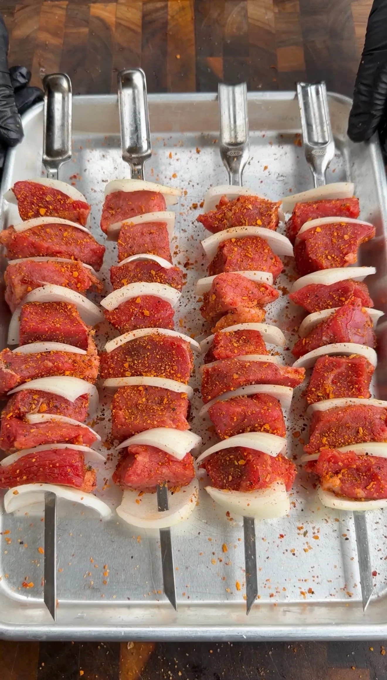 Raw cheese-crusted chimichurri steak kebabs with onions on metal skewers in aluminum tray ready for grilling