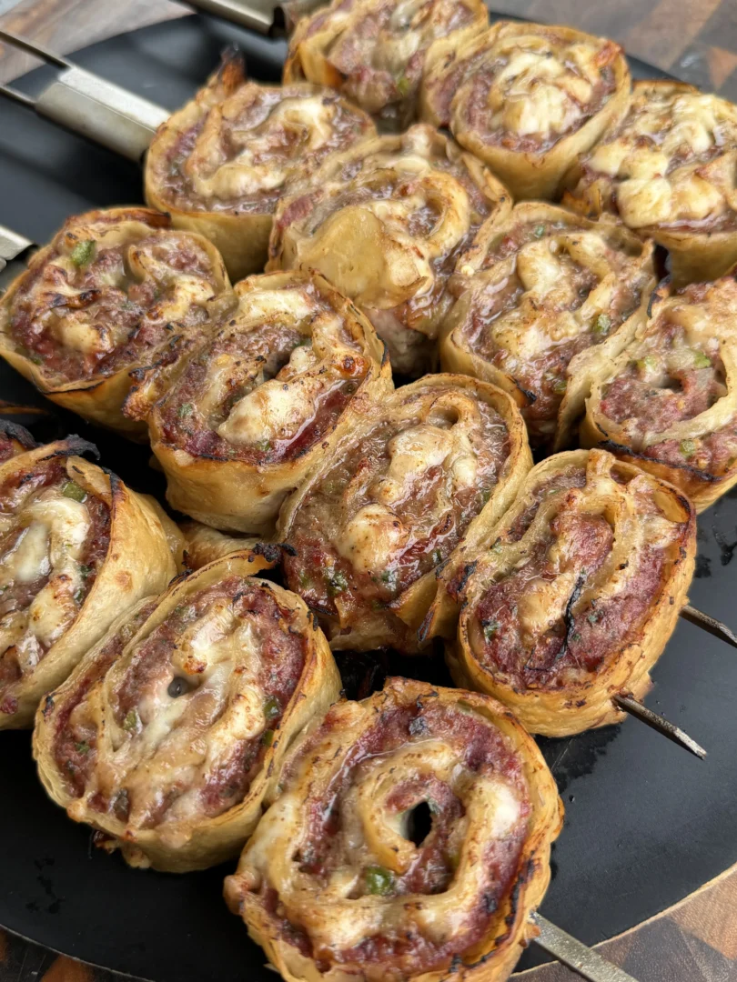 Golden-brown cheesy beef tortilla pinwheels threaded on metal skewers cooking on a black grill pan