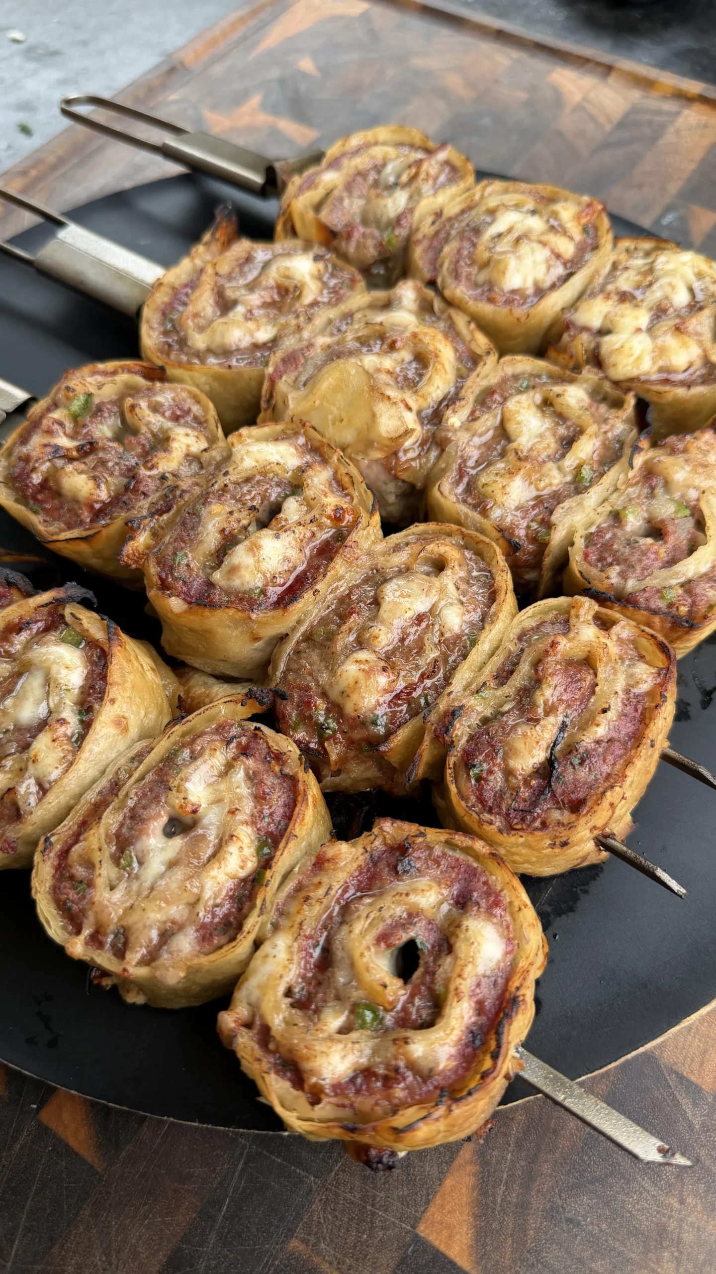 Golden-brown cheesy beef tortilla pinwheels threaded on metal skewers cooking on a black grill pan