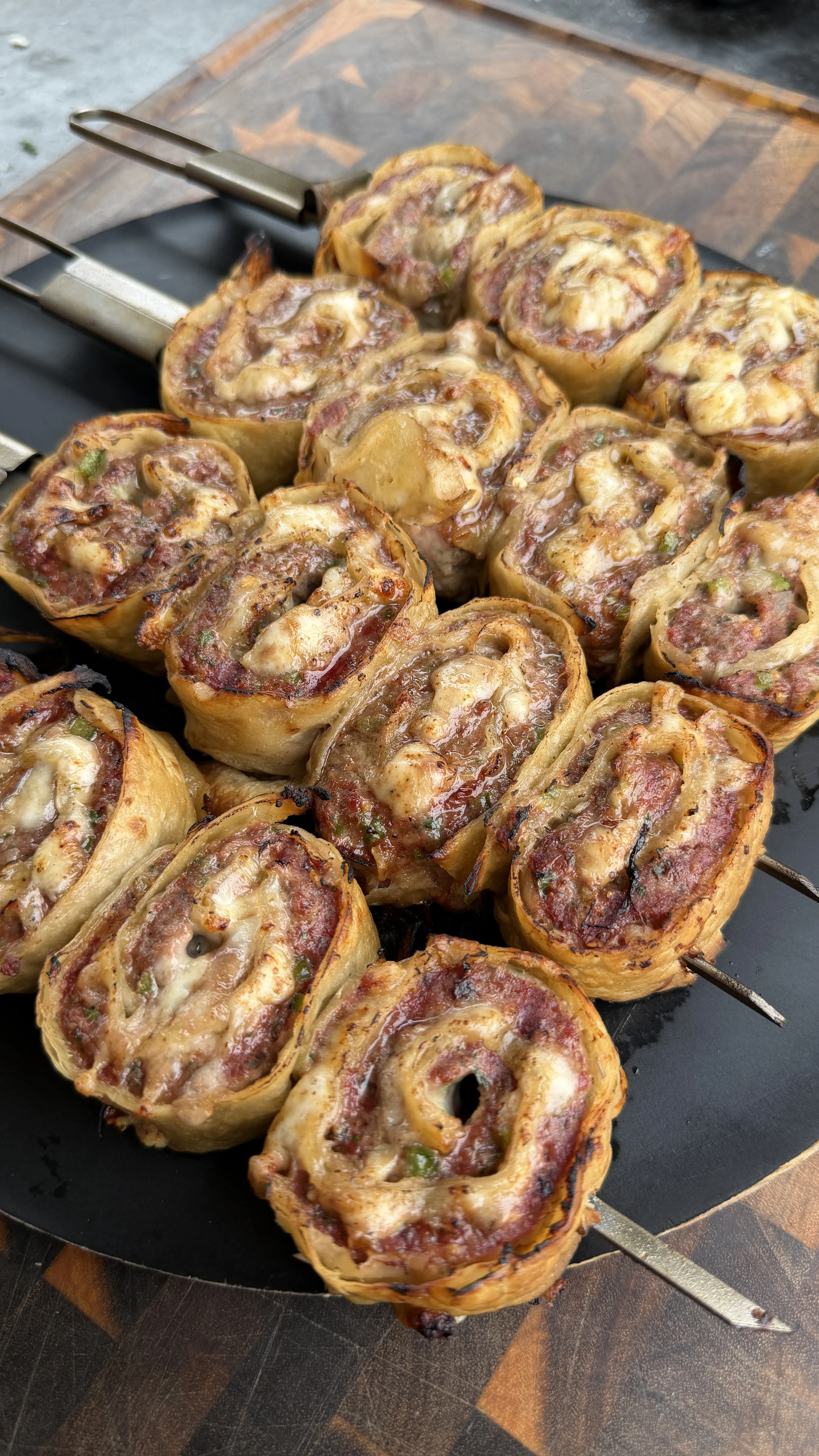 Golden-brown cheesy beef tortilla pinwheels threaded on metal skewers cooking on a black grill pan