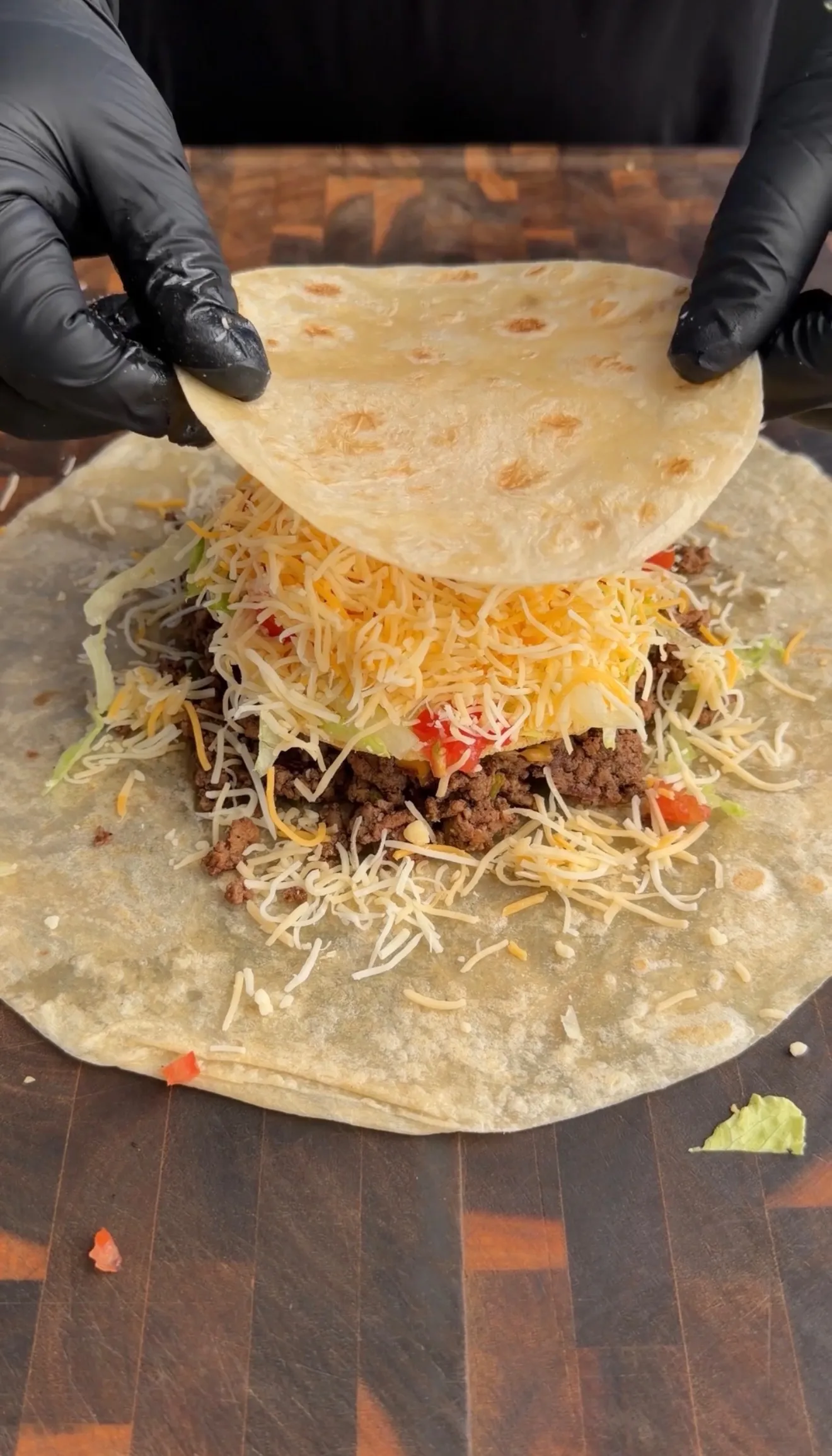 Hands in black gloves folding a flour tortilla over ground beef, cheese, and lettuce to assemble a classic crunchwrap