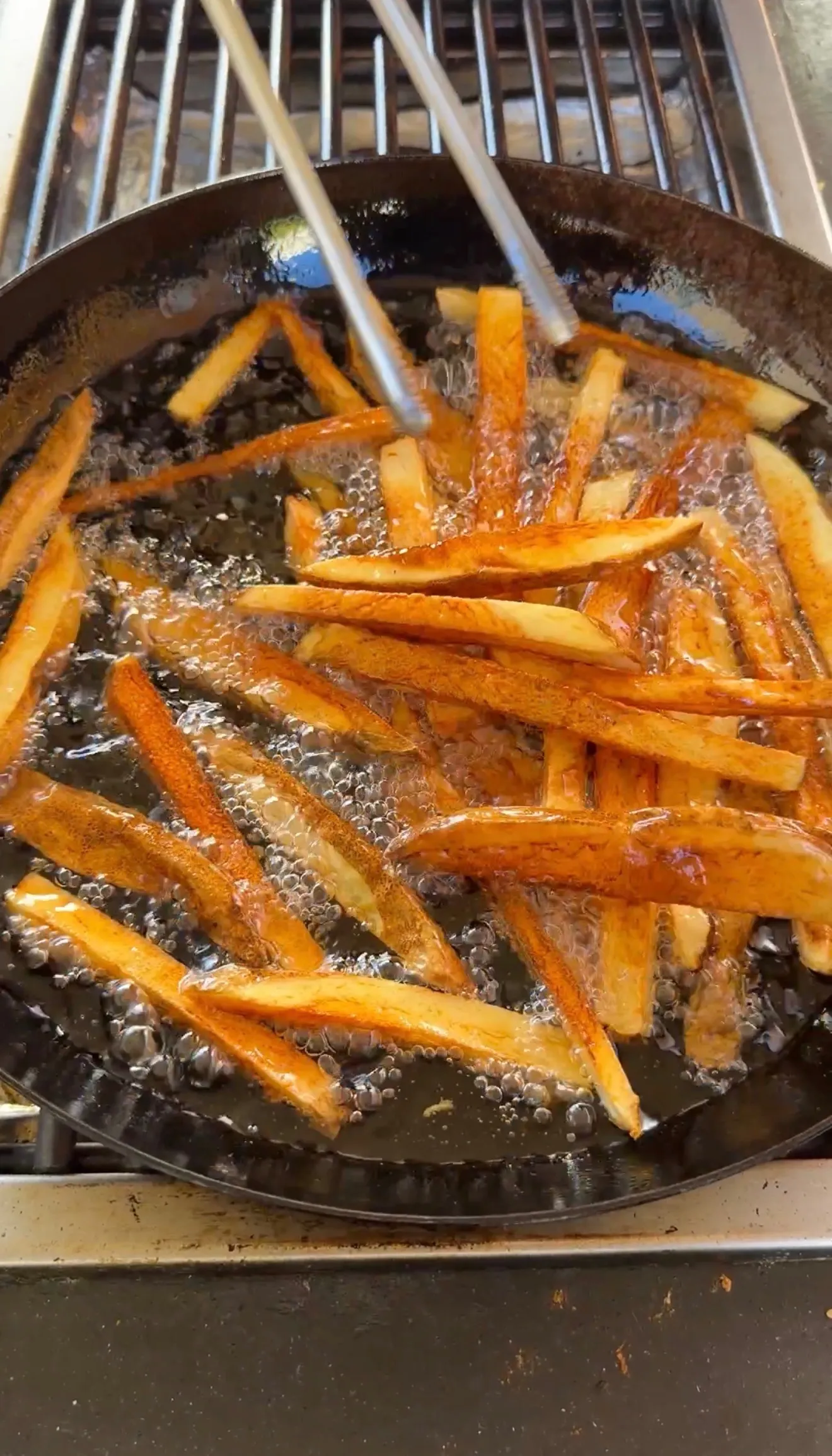 Golden french fries being deep fried in bubbling oil in a cast iron skillet with tongs