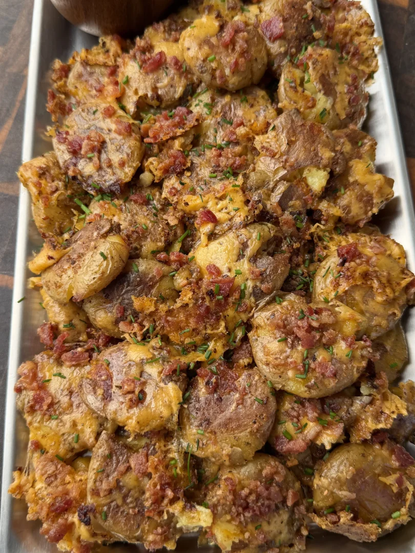 Crispy smashed mini potatoes topped with bacon bits and herbs on a metal tray with creamy dipping sauce