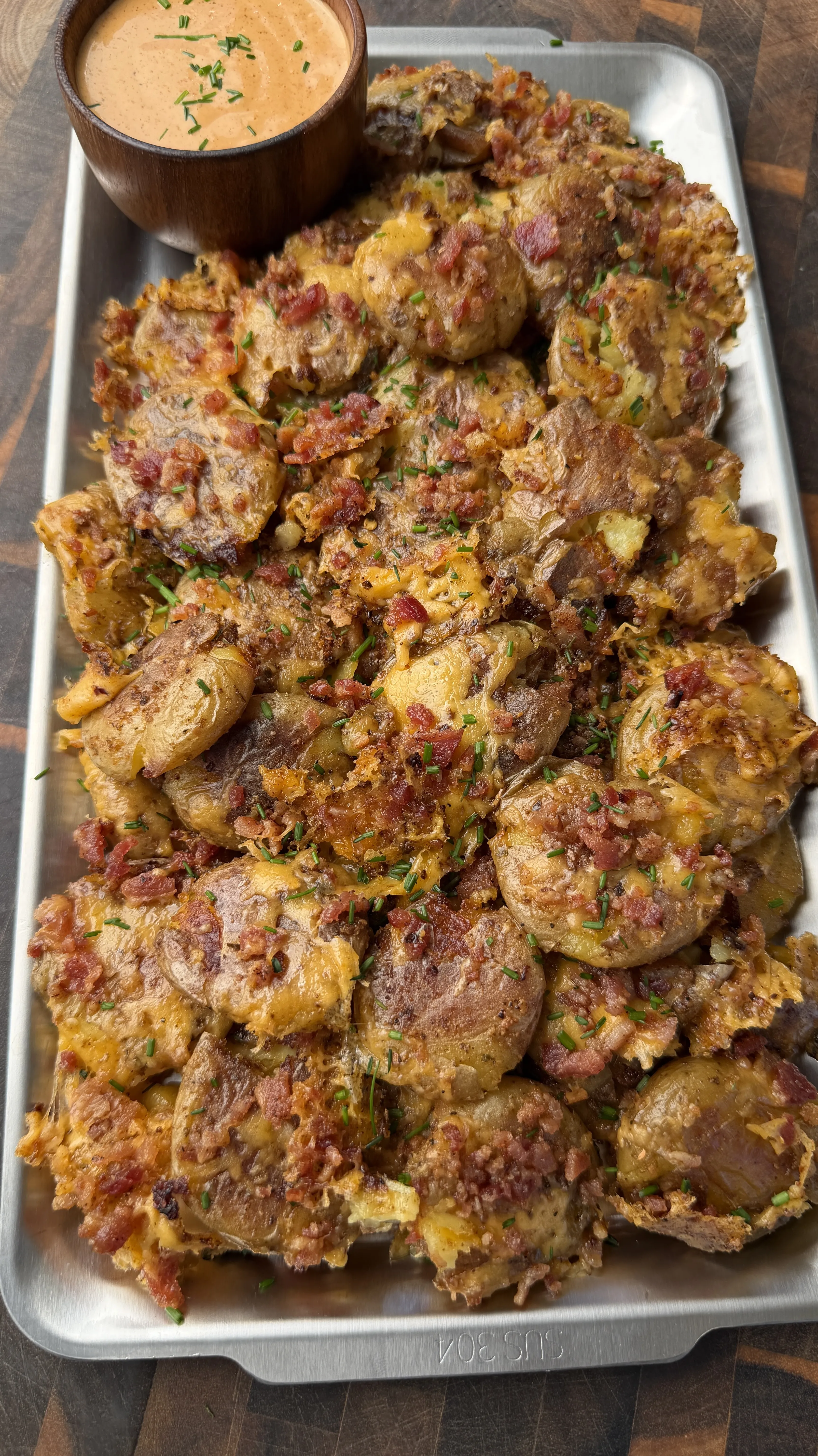 Crispy smashed mini potatoes topped with bacon bits and herbs on a metal tray with creamy dipping sauce