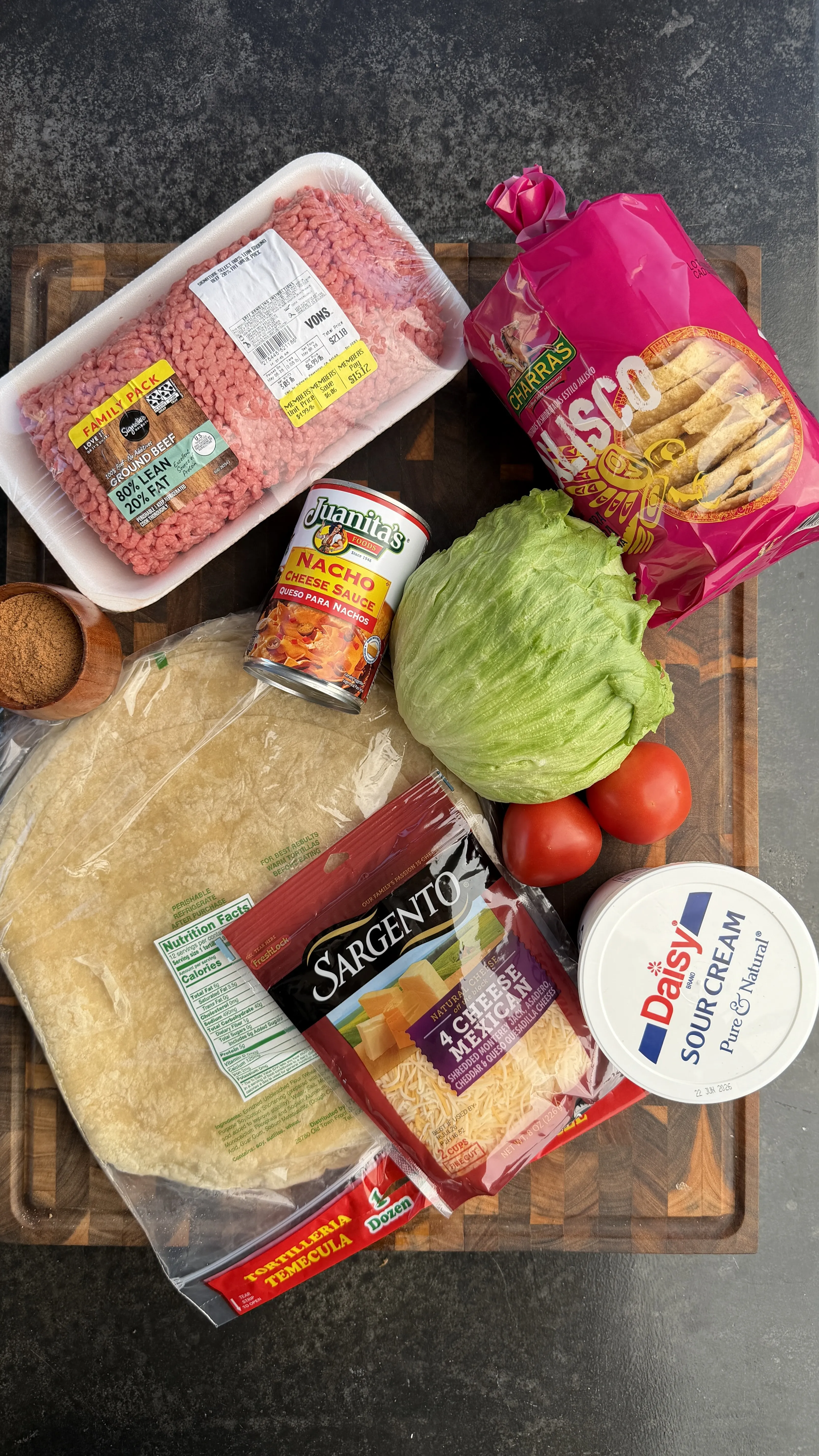 Ingredients for classic crunchwrap arranged on wooden cutting board including ground beef, tortillas, lettuce, tomatoes, cheese and sour cream