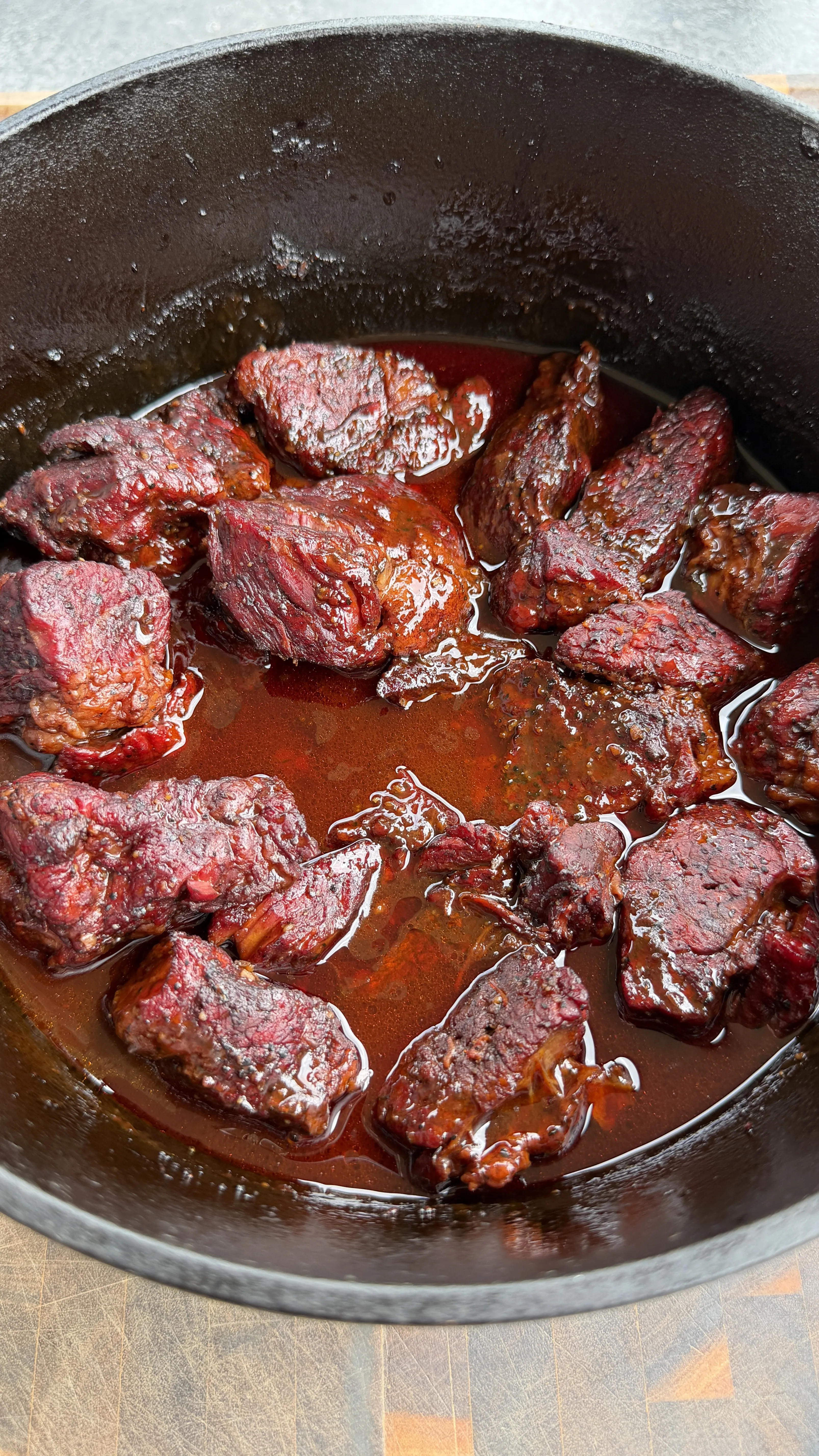 Beef chunks braising in rich Dr Pepper sauce in a black Dutch oven during cooking process