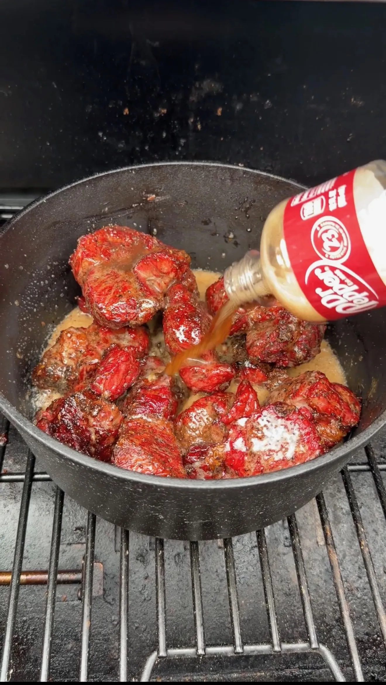 Beef chunks cooking in cast iron pan with Dr Pepper being poured over them for pulled beef sandwiches