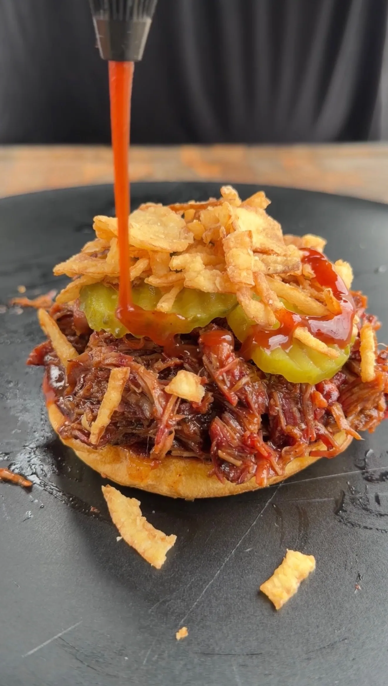 Dr Pepper pulled beef sandwich topped with pickles and crispy onions, drizzled with hot sauce on dark slate plate