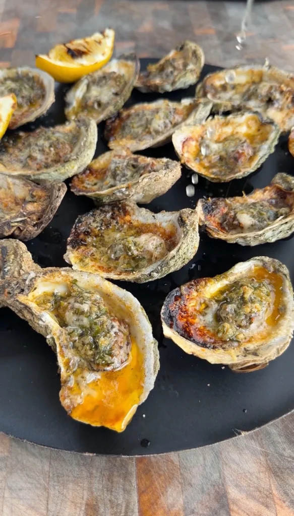 Garlic herb Parmesan oysters grilling on a black grate with melted cheese and herbs, lemon wedge visible