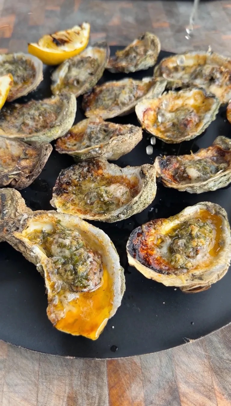 Garlic herb Parmesan oysters grilling on a black grate with melted cheese and herbs, lemon wedge visible