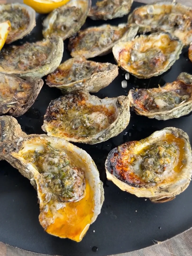 Garlic herb Parmesan oysters grilling on a black grate with melted cheese and herbs, lemon wedge visible