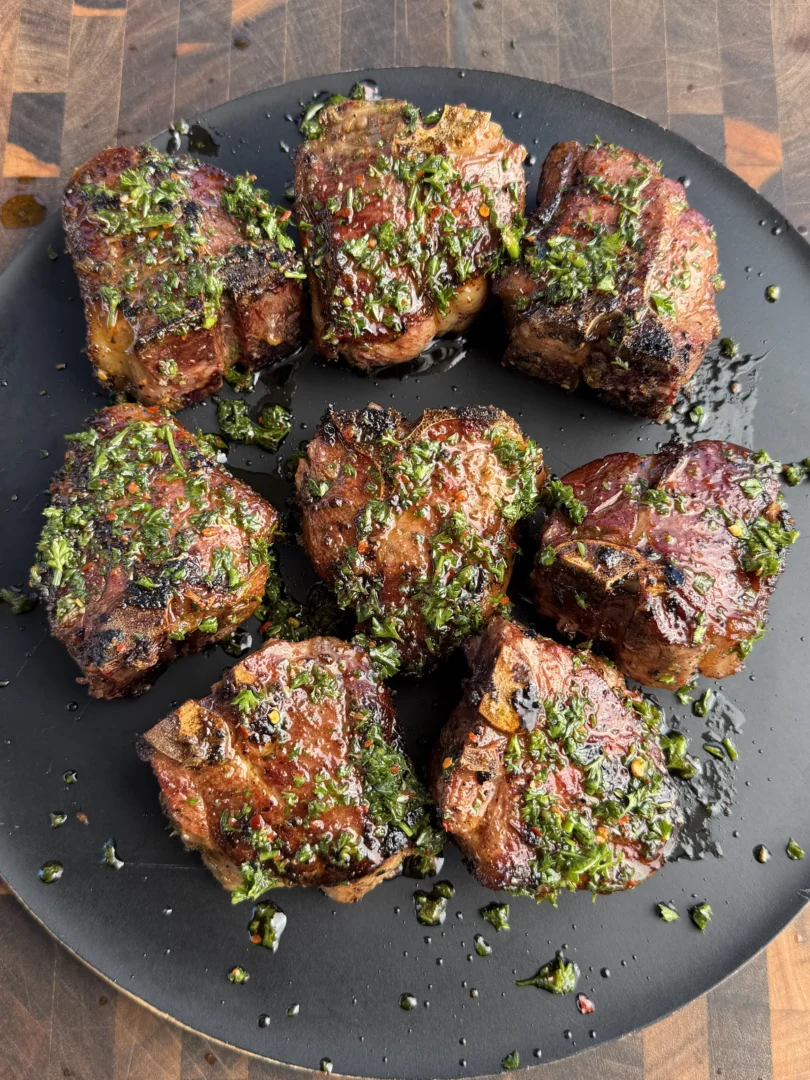 Eight grilled lamb loin chops topped with fresh green herb sauce arranged on a dark round plate