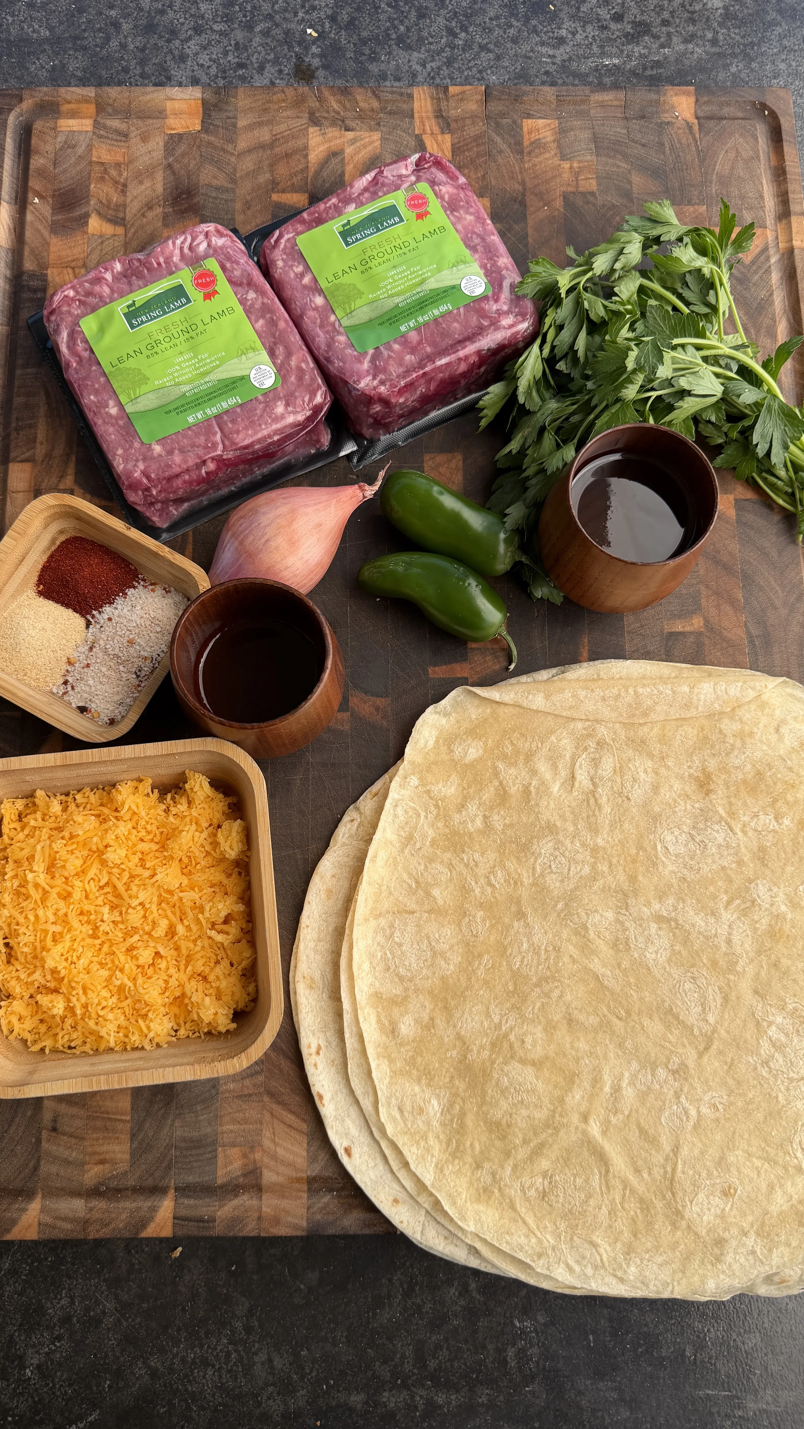 Raw ground lamb, flour tortillas, shredded cheese, jalapenos, herbs and spices arranged on wooden cutting board