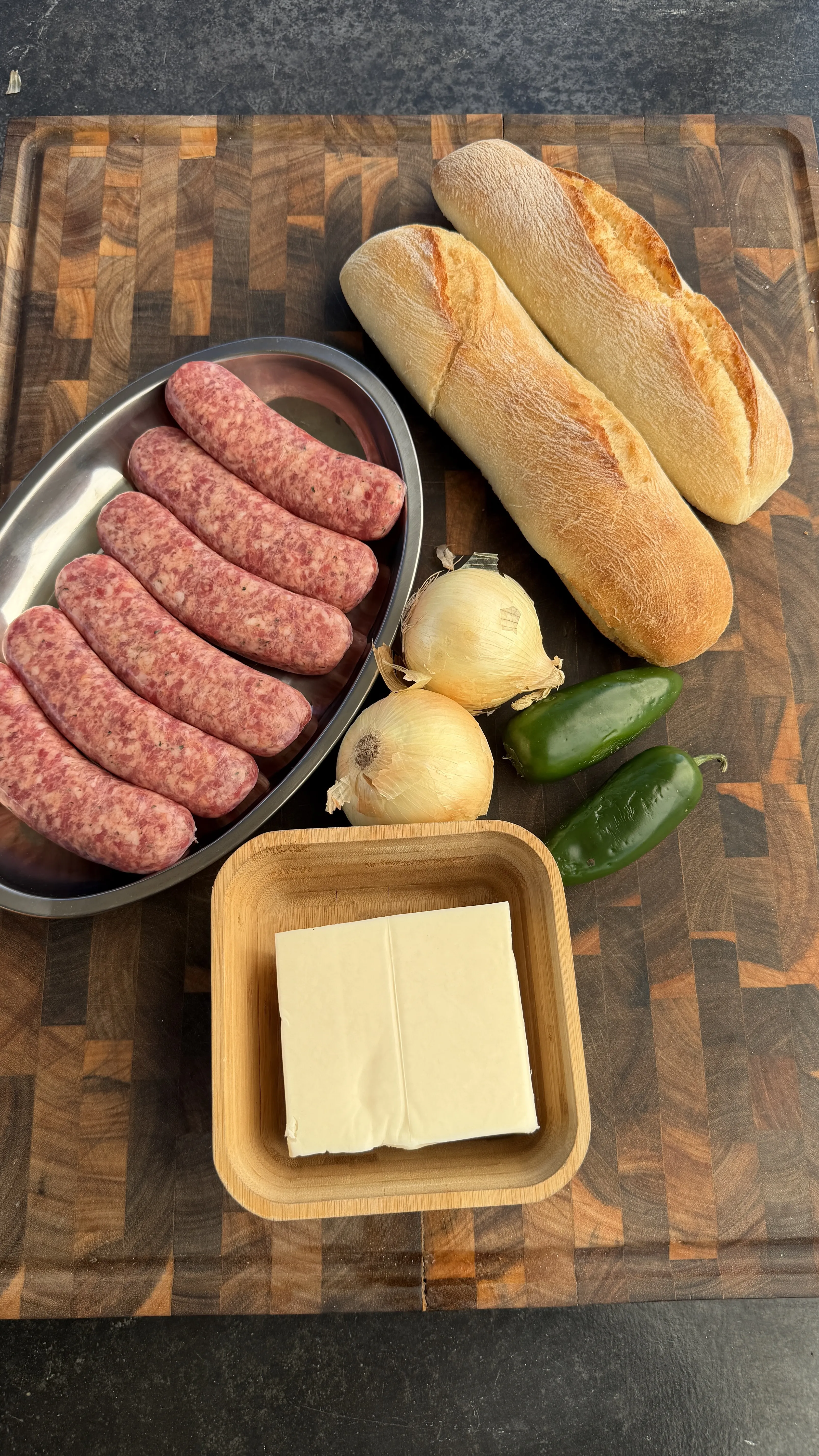 Raw sausages, bread rolls, cheese, onions and peppers arranged on wooden cutting board for stuffed bread boats