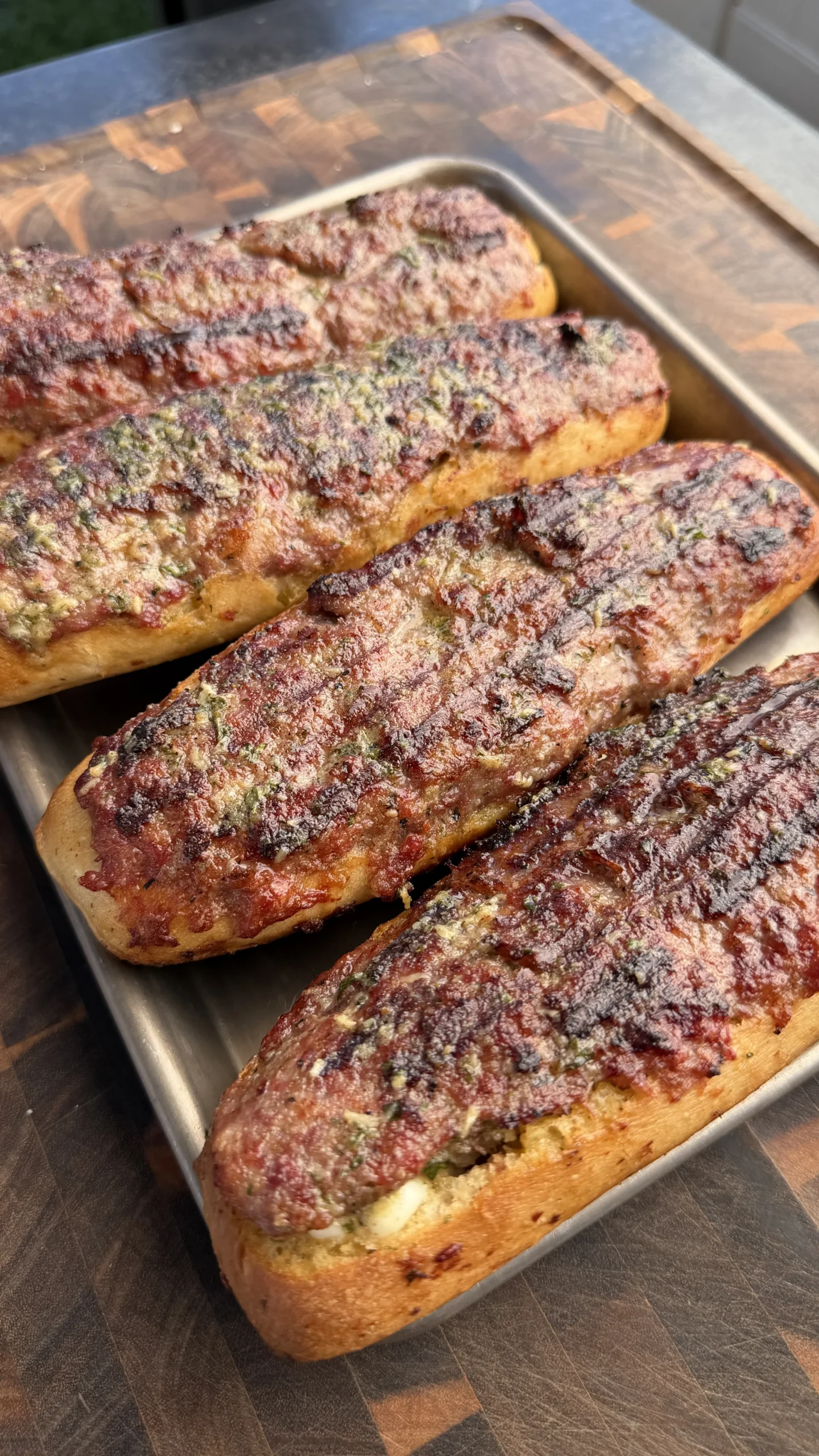 Four grilled sausage stuffed bread boats with melted cheese and herbs on a metal baking tray