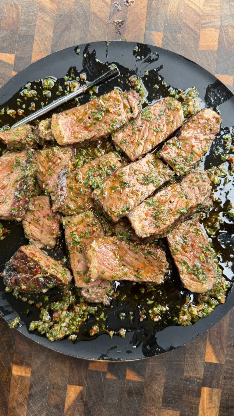 Grilled strip steaks topped with green herb board sauce in a dark skillet with tongs on wooden cutting board