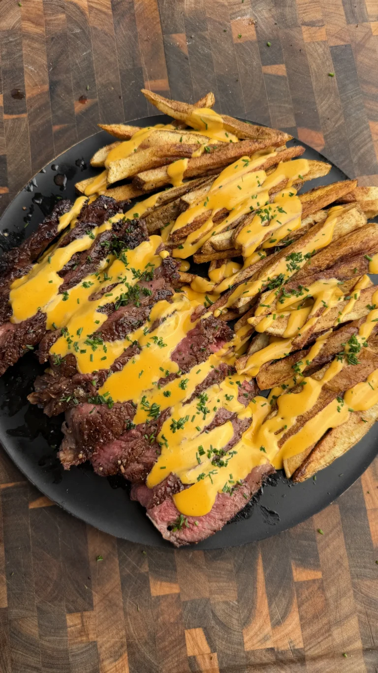 Sliced grilled tomahawk steak with crispy potato fries drizzled in golden cheese sauce on black plate