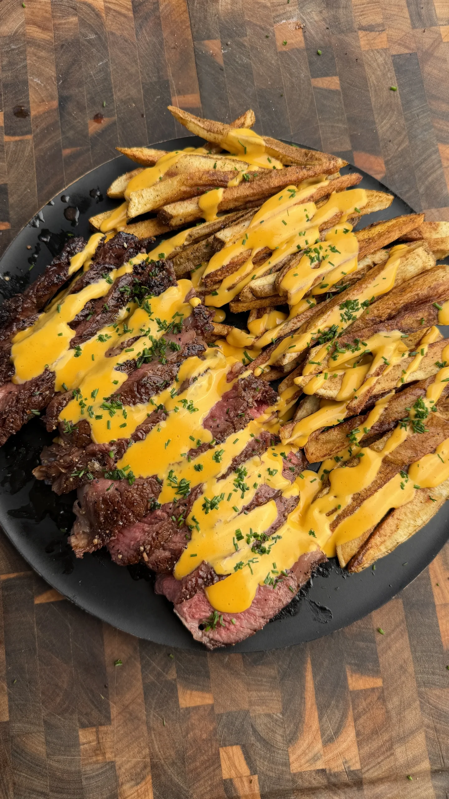 Sliced grilled tomahawk steak with crispy potato fries drizzled in golden cheese sauce on black plate
