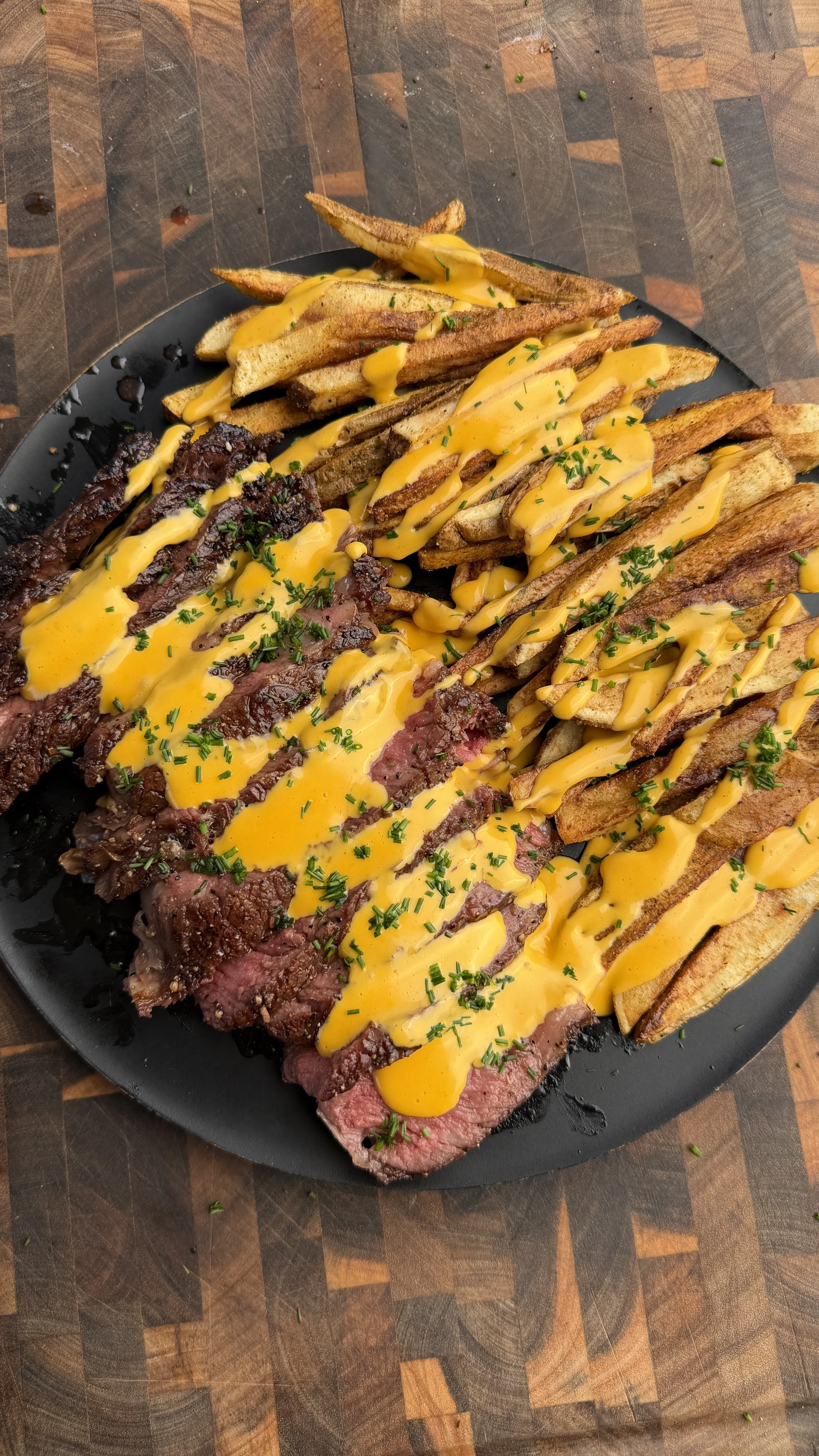 Sliced grilled tomahawk steak with crispy potato fries drizzled in golden cheese sauce on black plate