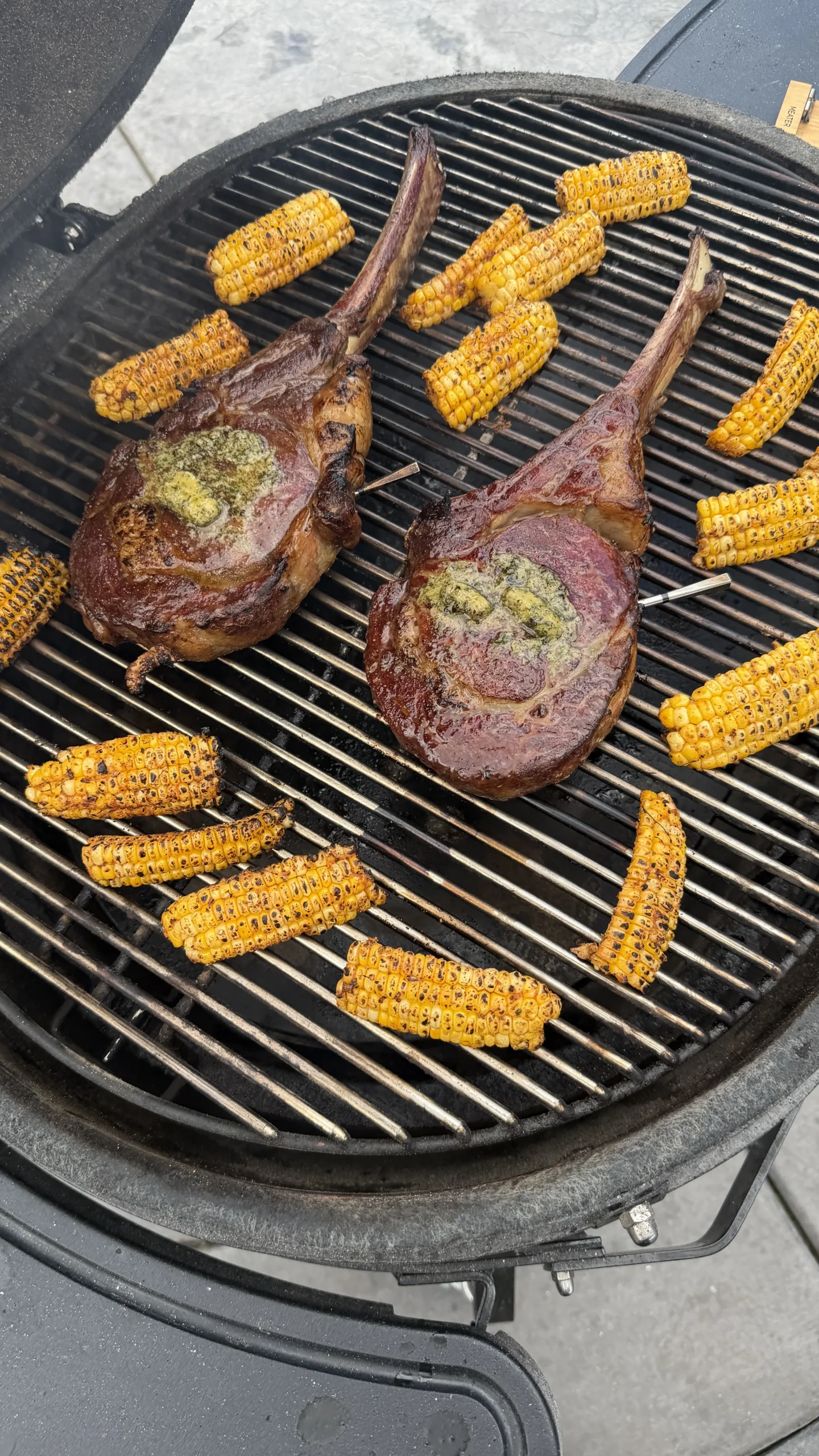 Two tomahawk steaks with herb butter and grilled mini corn ribs cooking on a round charcoal grill