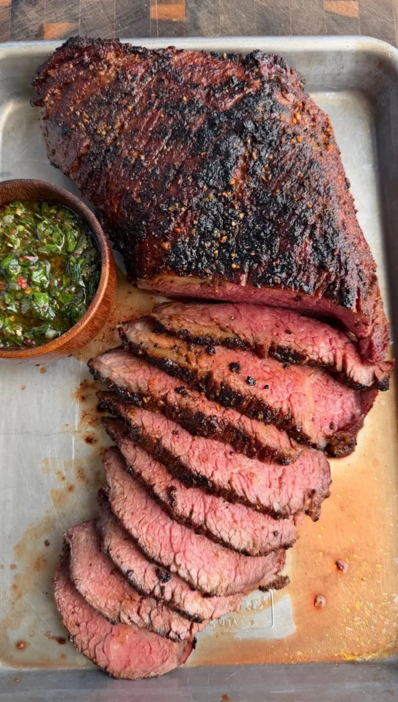 Sliced grilled tri-tip roast with charred crust on cutting board served with green onion chimichurri sauce