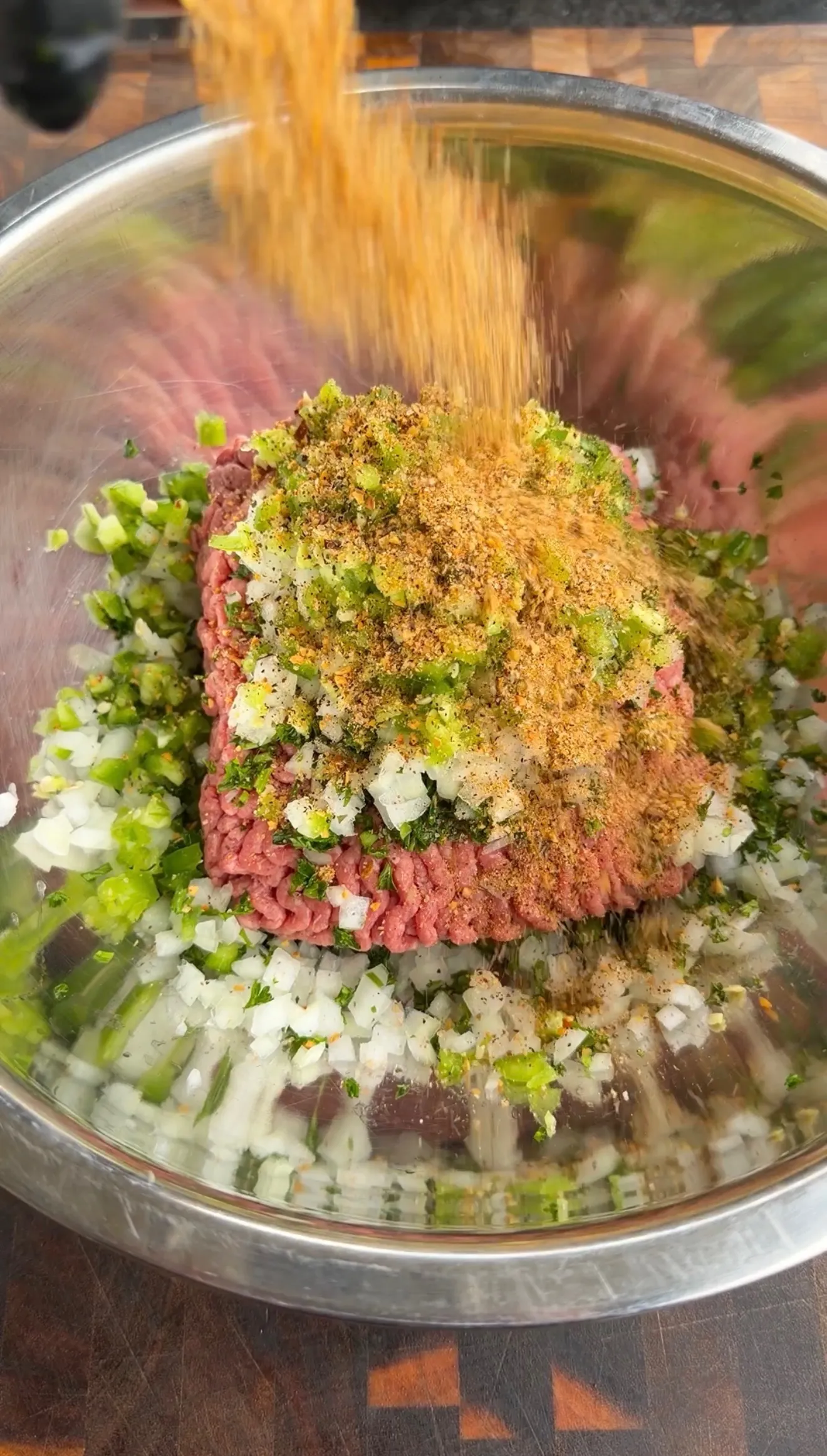 Raw ground beef mixed with diced onions, herbs and golden seasoning spices in a stainless steel mixing bowl