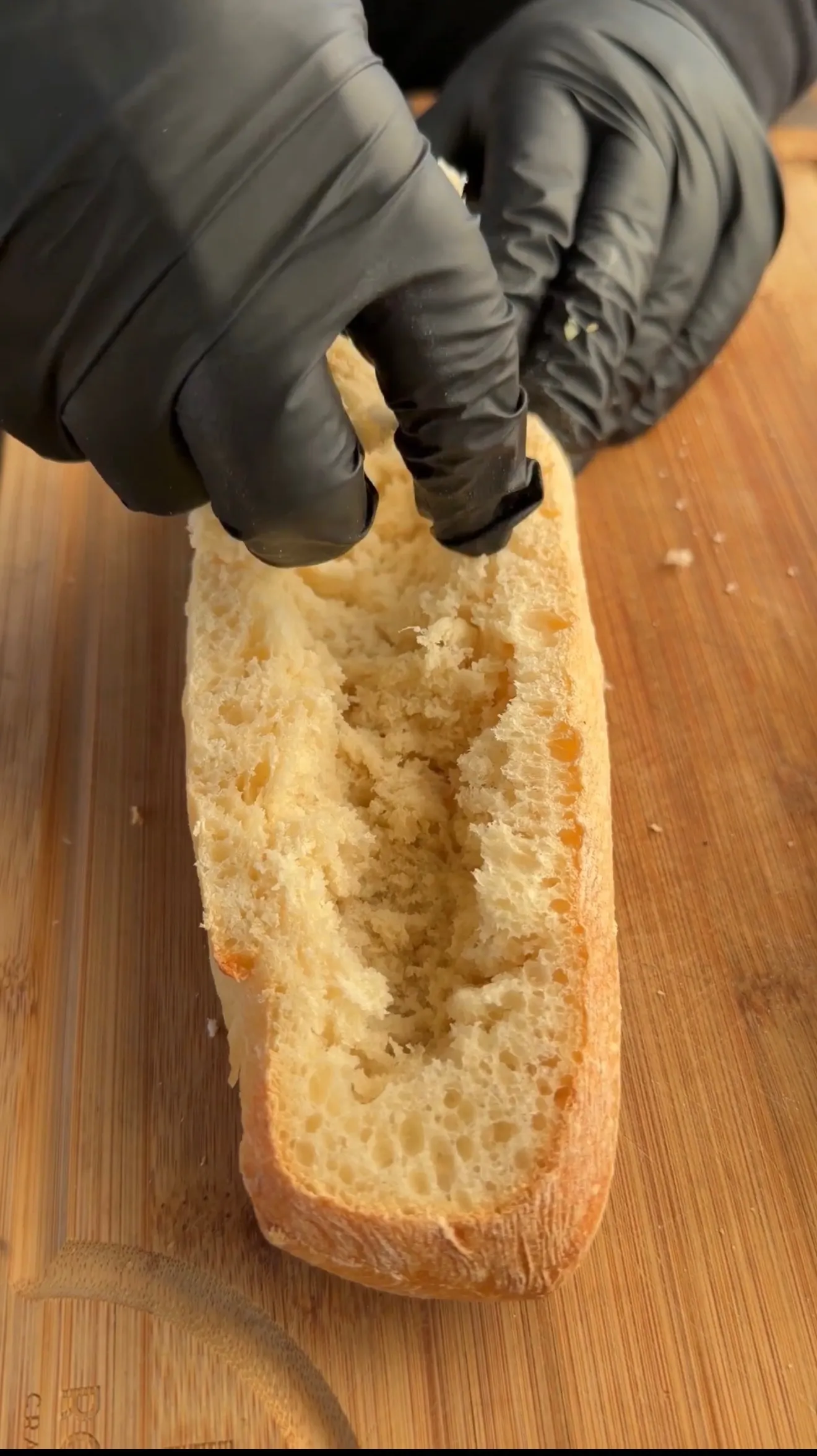 Hands in black gloves hollowing out a crusty bread loaf on wooden cutting board to make stuffed bread boat
