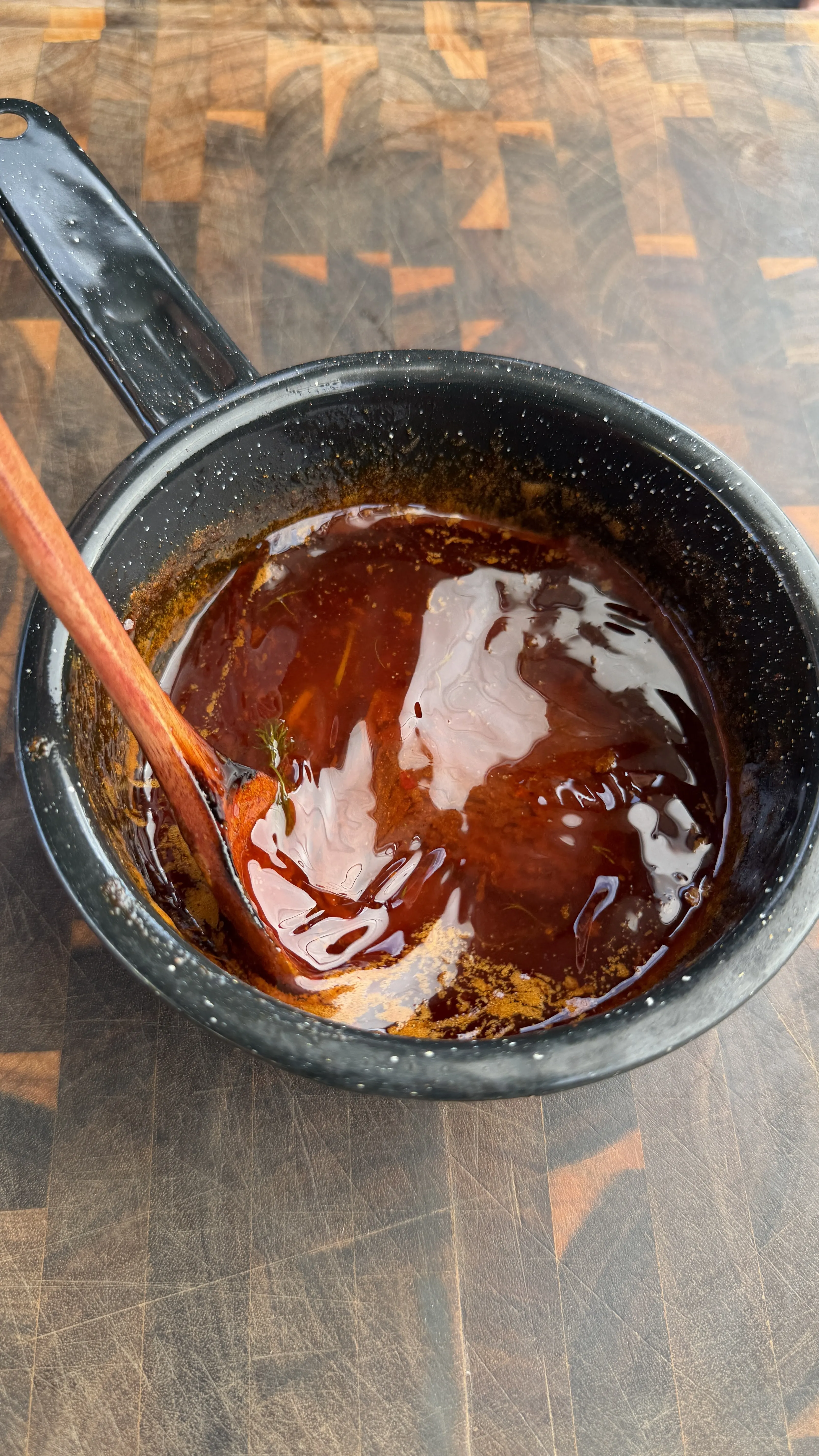 Dark red Nashville hot sauce being stirred with wooden spoon in black speckled saucepan