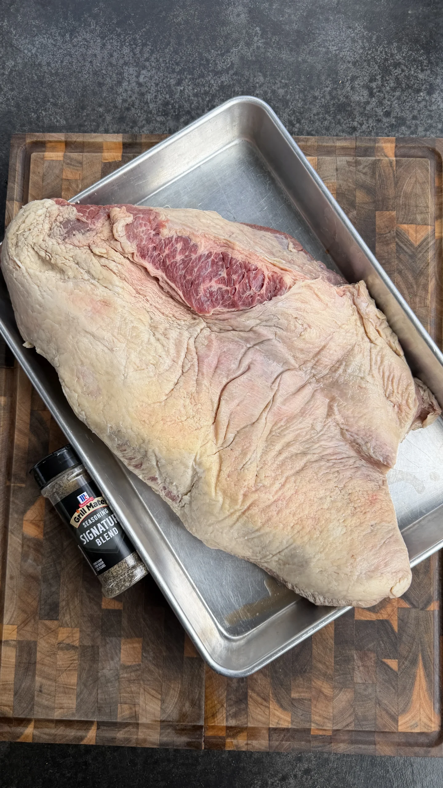 Raw beef brisket in metal roasting pan on wooden cutting board with seasoning bottle for barbecue smoking