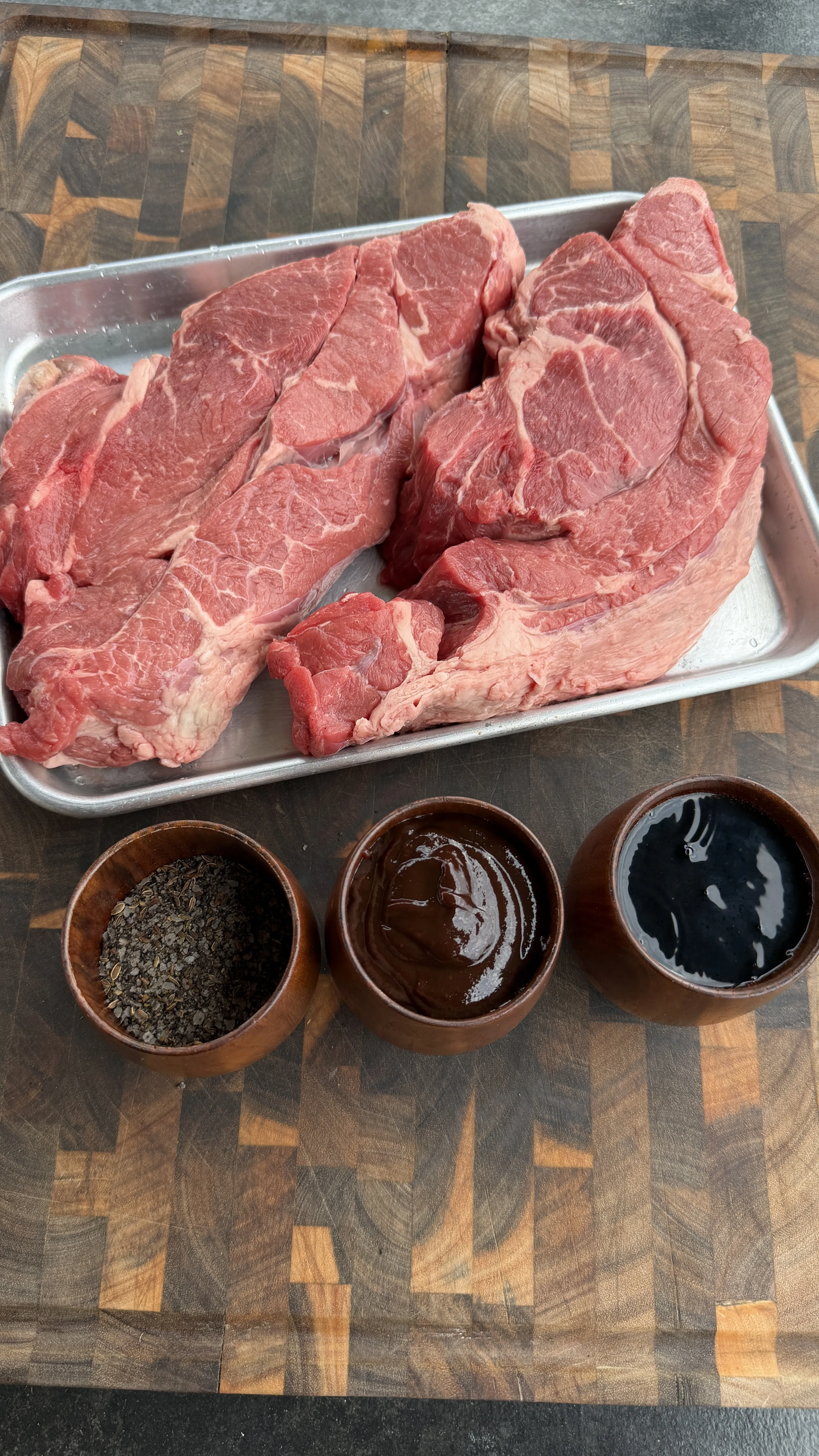 Raw beef chuck roast in metal tray with three bowls of seasonings and Dr Pepper marinade on cutting board