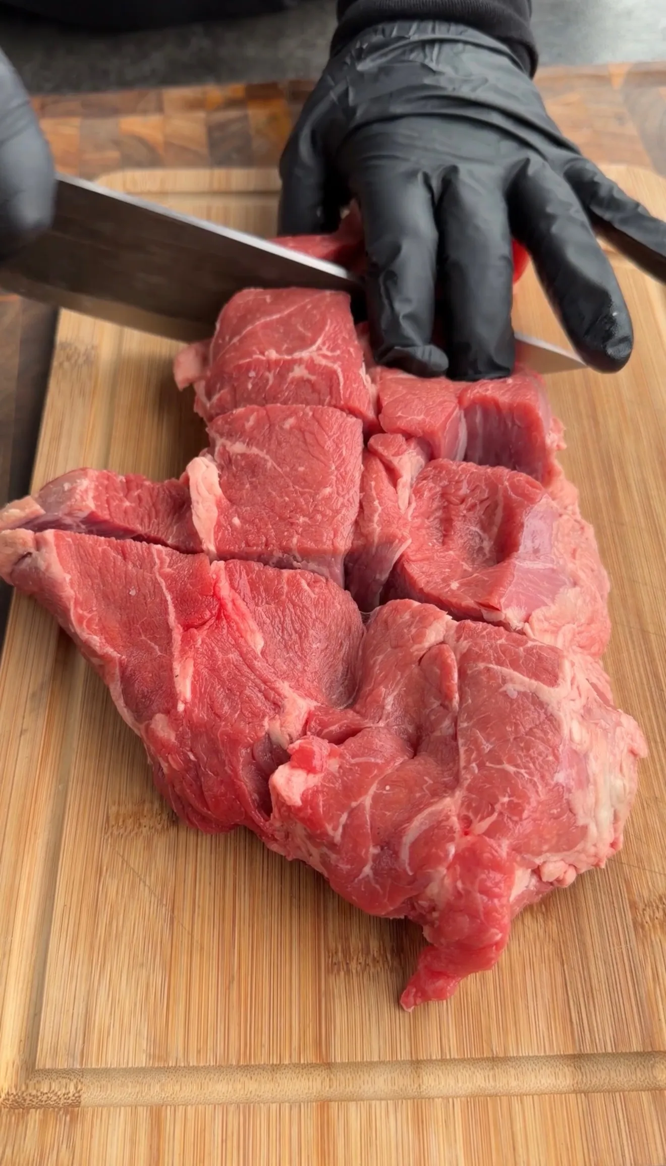 Raw beef chunks being cut with knife on wooden cutting board, person wearing black gloves for Dr Pepper pulled beef