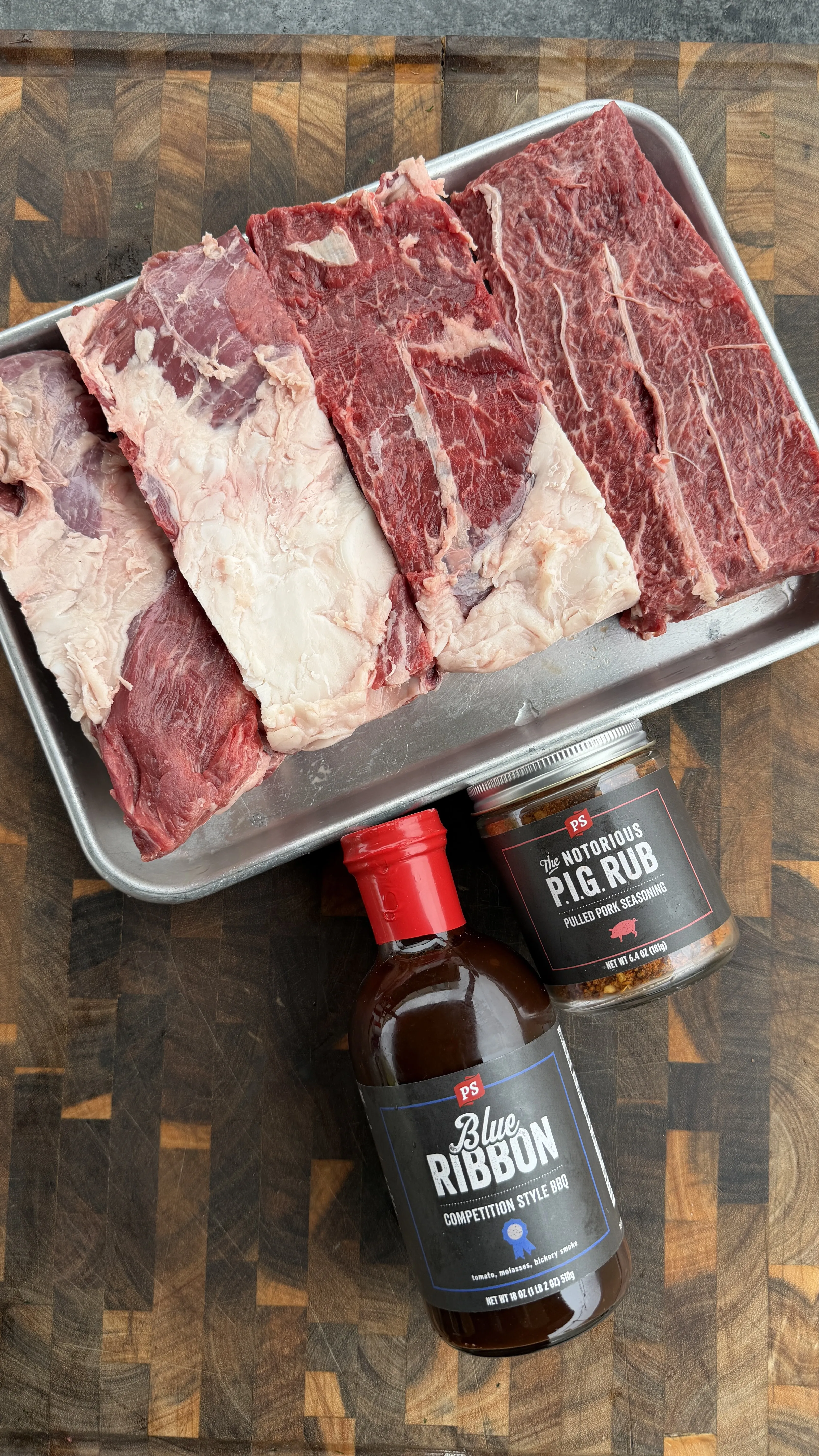Raw beef ribs on metal tray with barbecue sauce and seasoning on wooden cutting board