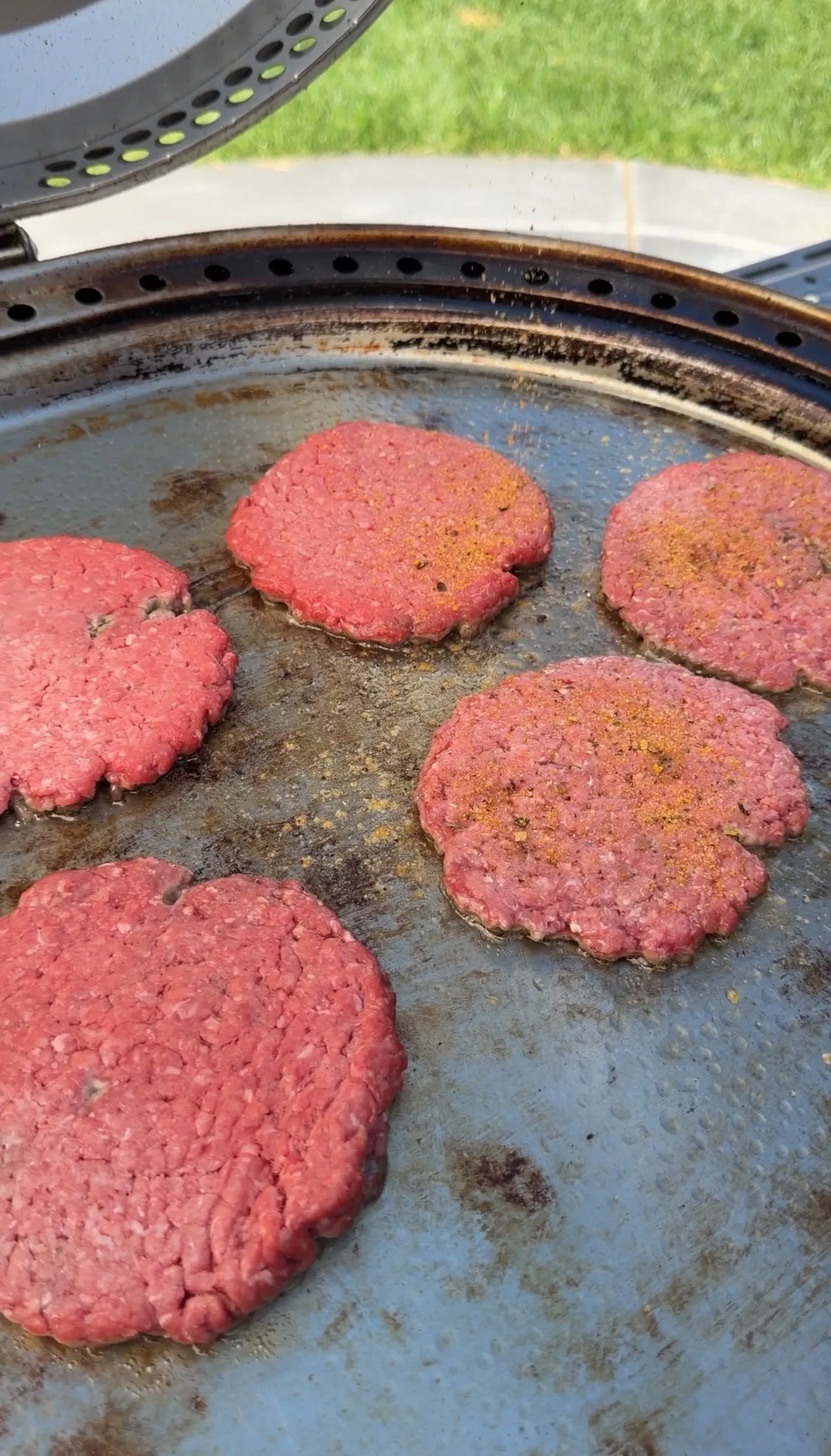 Six raw ground beef burger patties with seasoning cooking on a round outdoor griddle