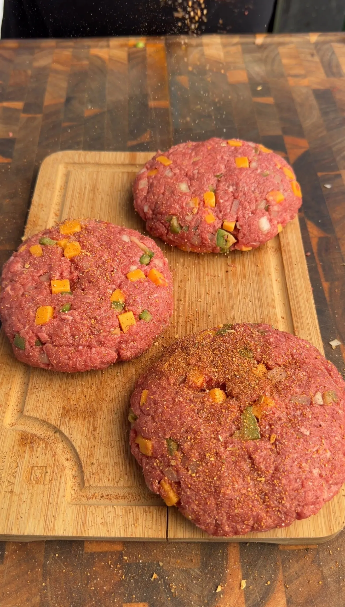 Three raw smoked cheddar jalapeño stuffed burger patties with diced peppers on wooden cutting board